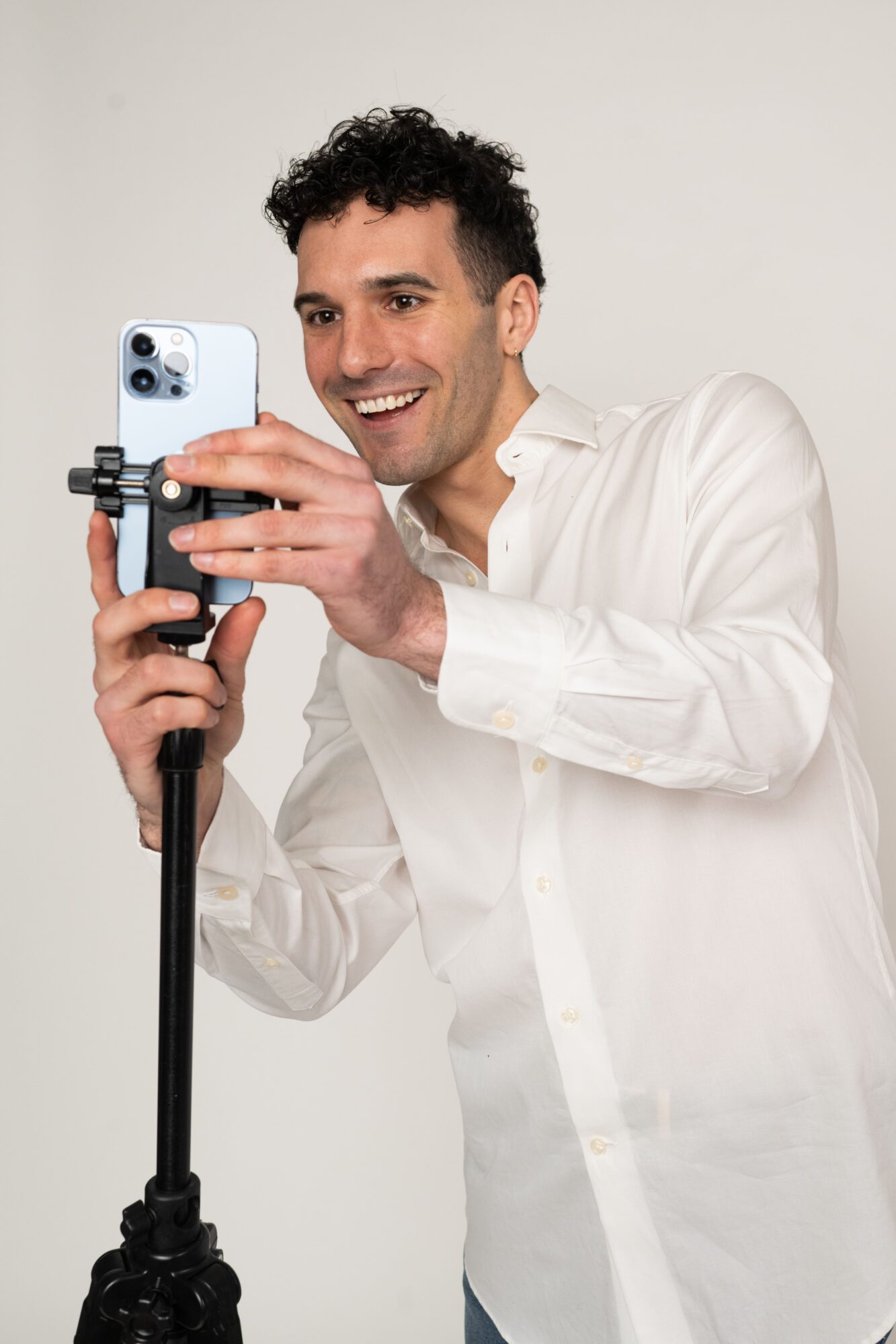 Man with dark curly hair smiling and taking a photo with a smartphone on a tripod, wearing a white shirt.