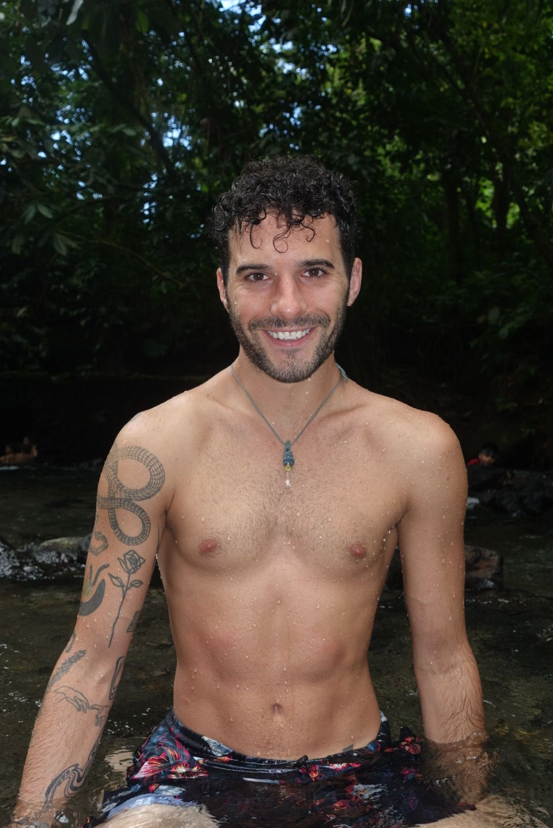 A shirtless man with tattoos on his arm, smiling outdoors with trees in the background.