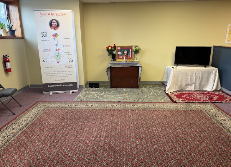 Room corner with a poster, a table with flowers and a framed photo, and a TV on a table, with a large carpet.