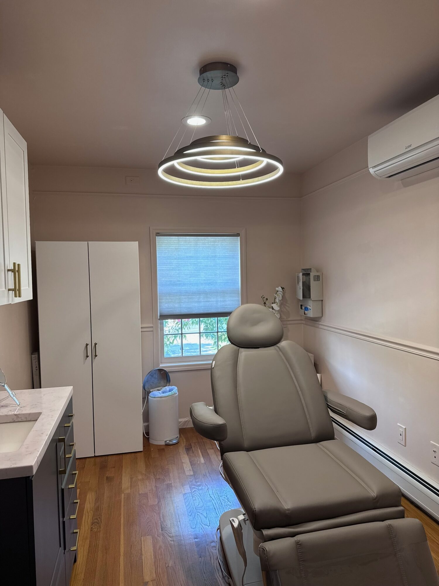 Medical examination room with adjustable chair, white cabinet, window with blinds, and ceiling light fixture.