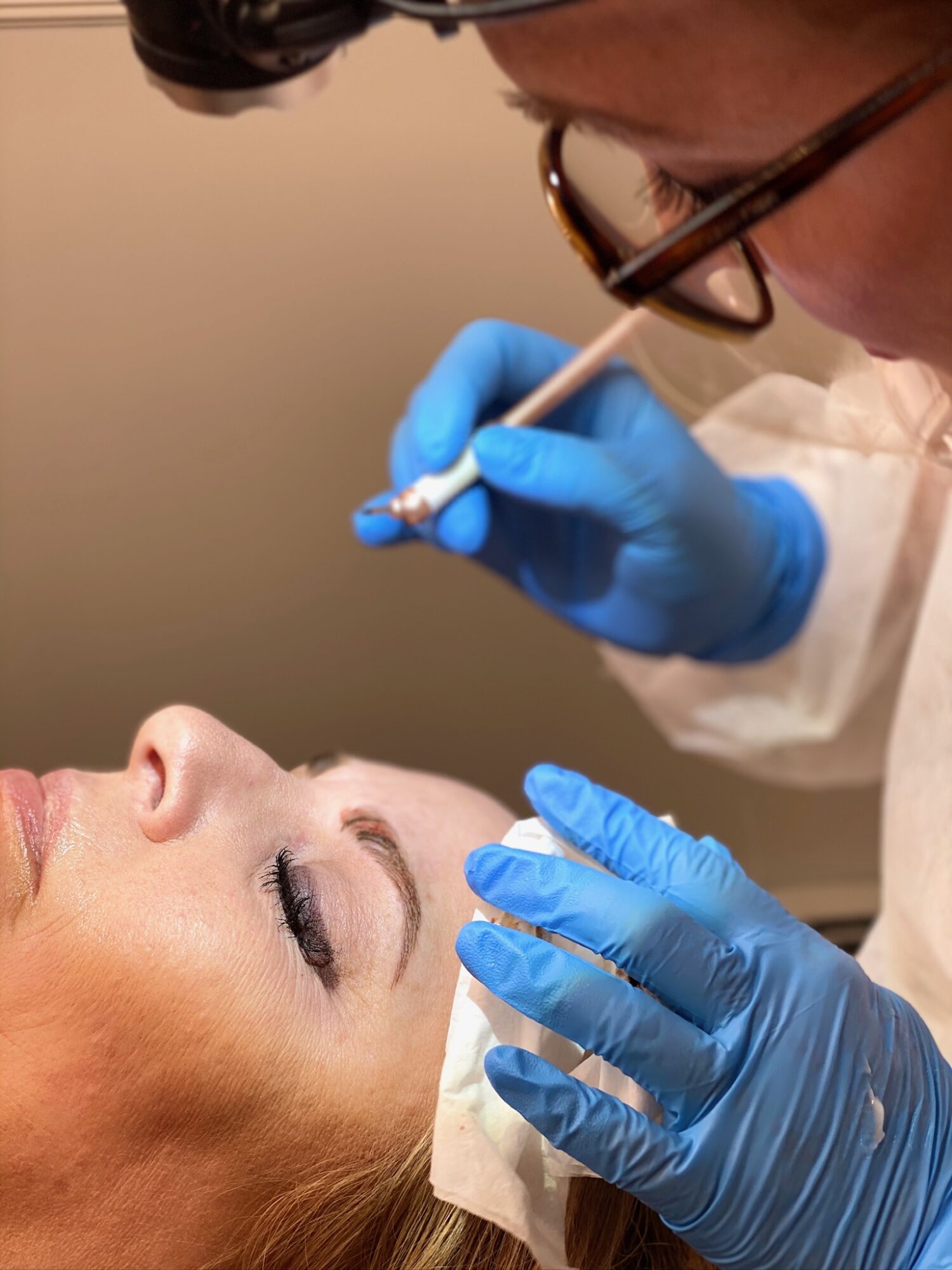 A person receiving a cosmetic treatment, lying down with eyes closed, while a professional in blue gloves applies a procedure near the eye.