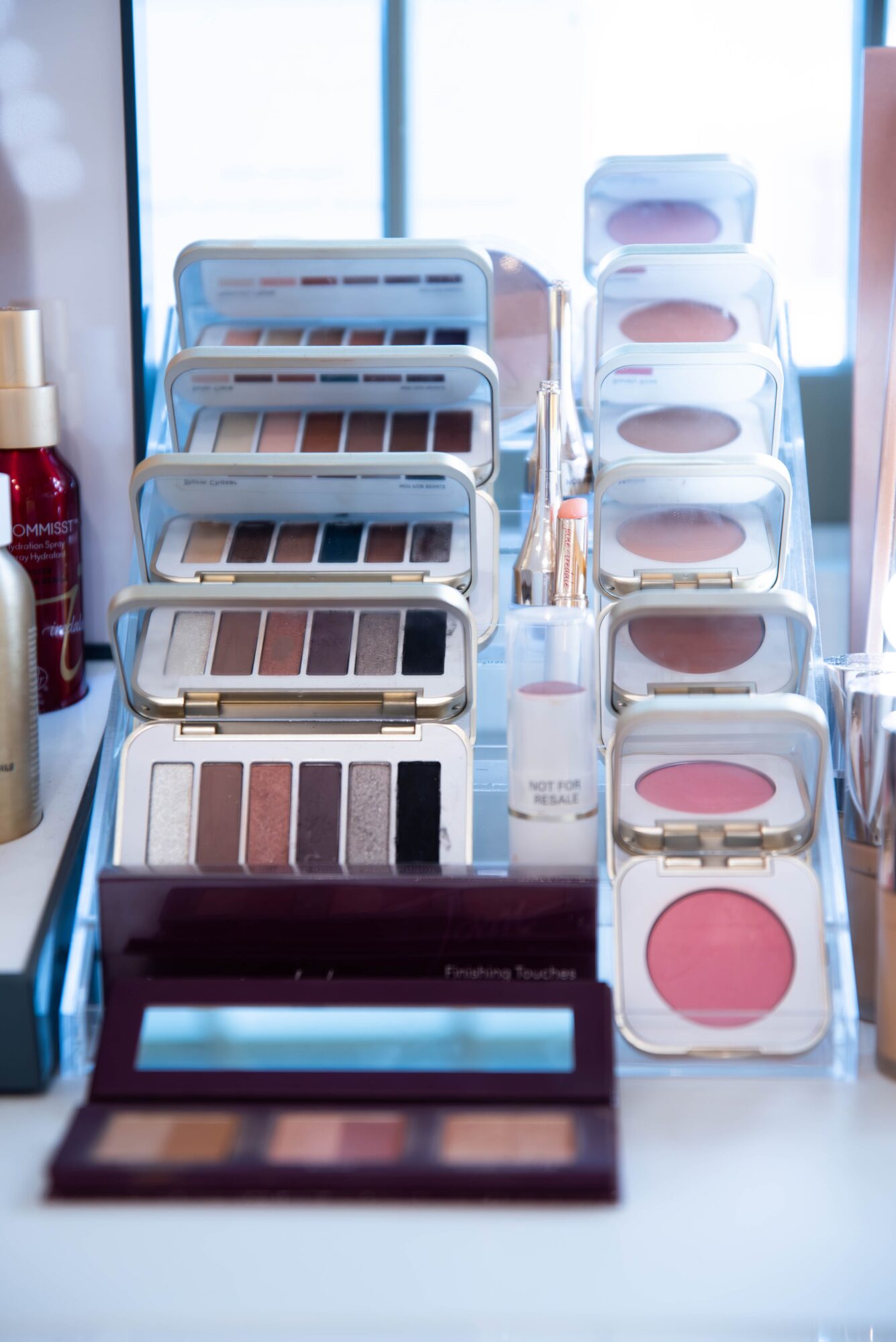 Makeup products including eyeshadow palettes, blushes, and a compact on a white surface with a window in the background.