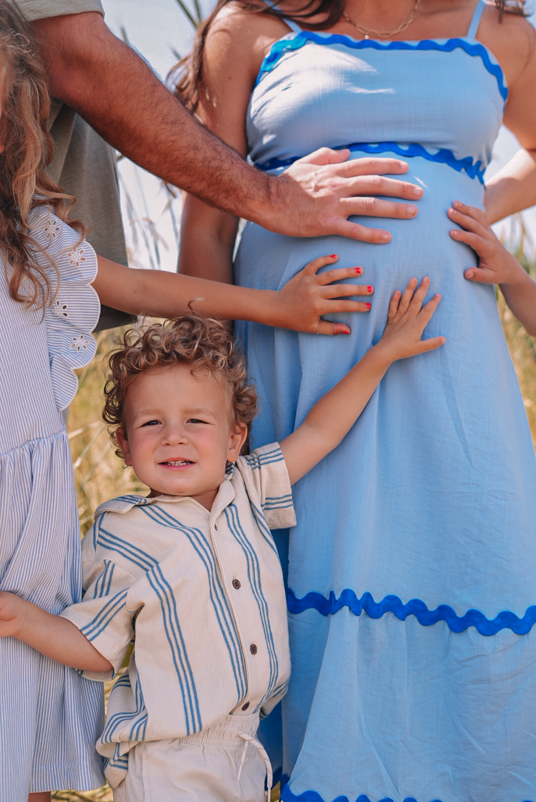 Child with curly hair smiling and reaching out, standing next to a woman in a blue dress, with other people partially visible.
