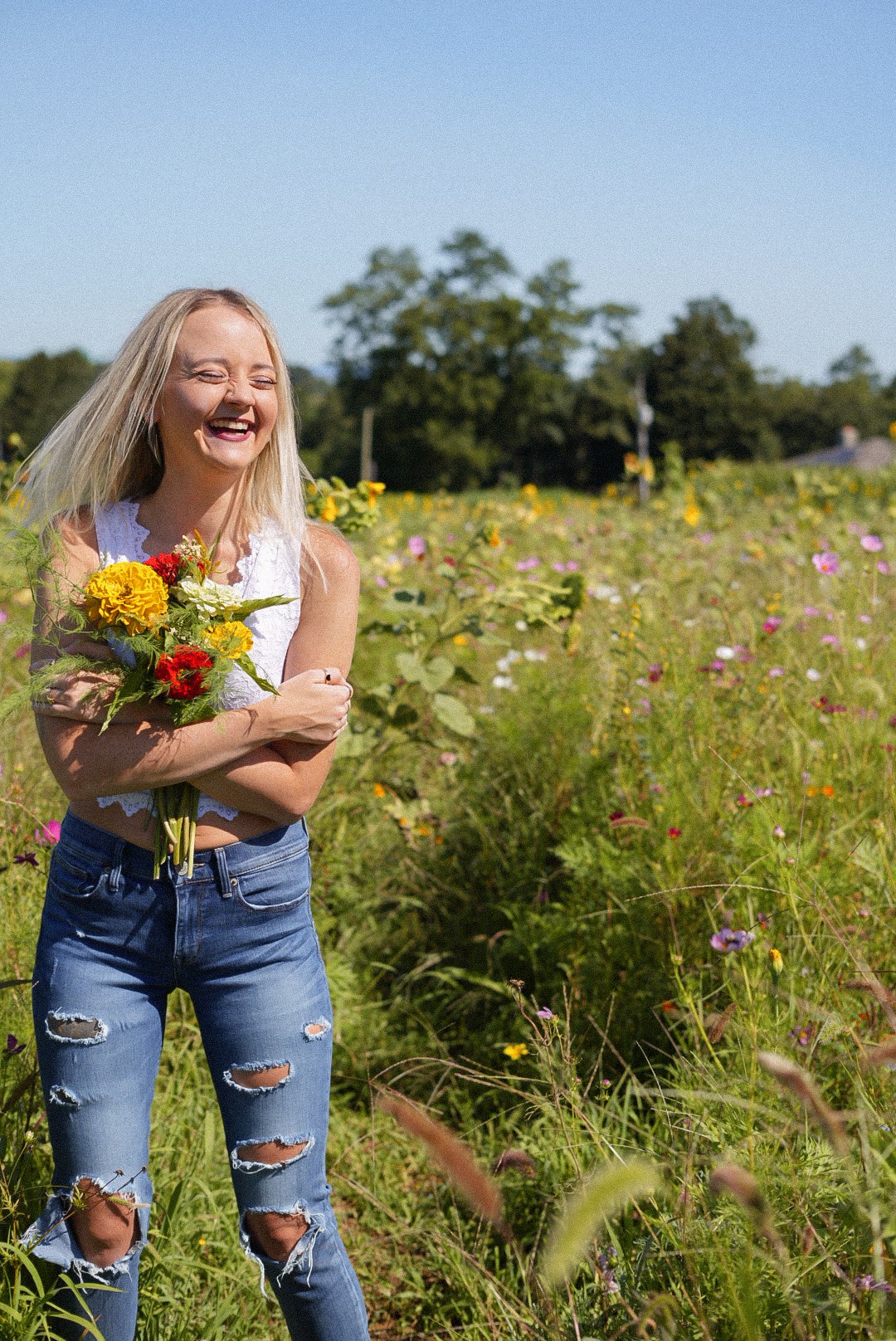 Smiling woman holding a bouquet of flowers in a field with green plants and colorful flowers, under a clear blue sky.