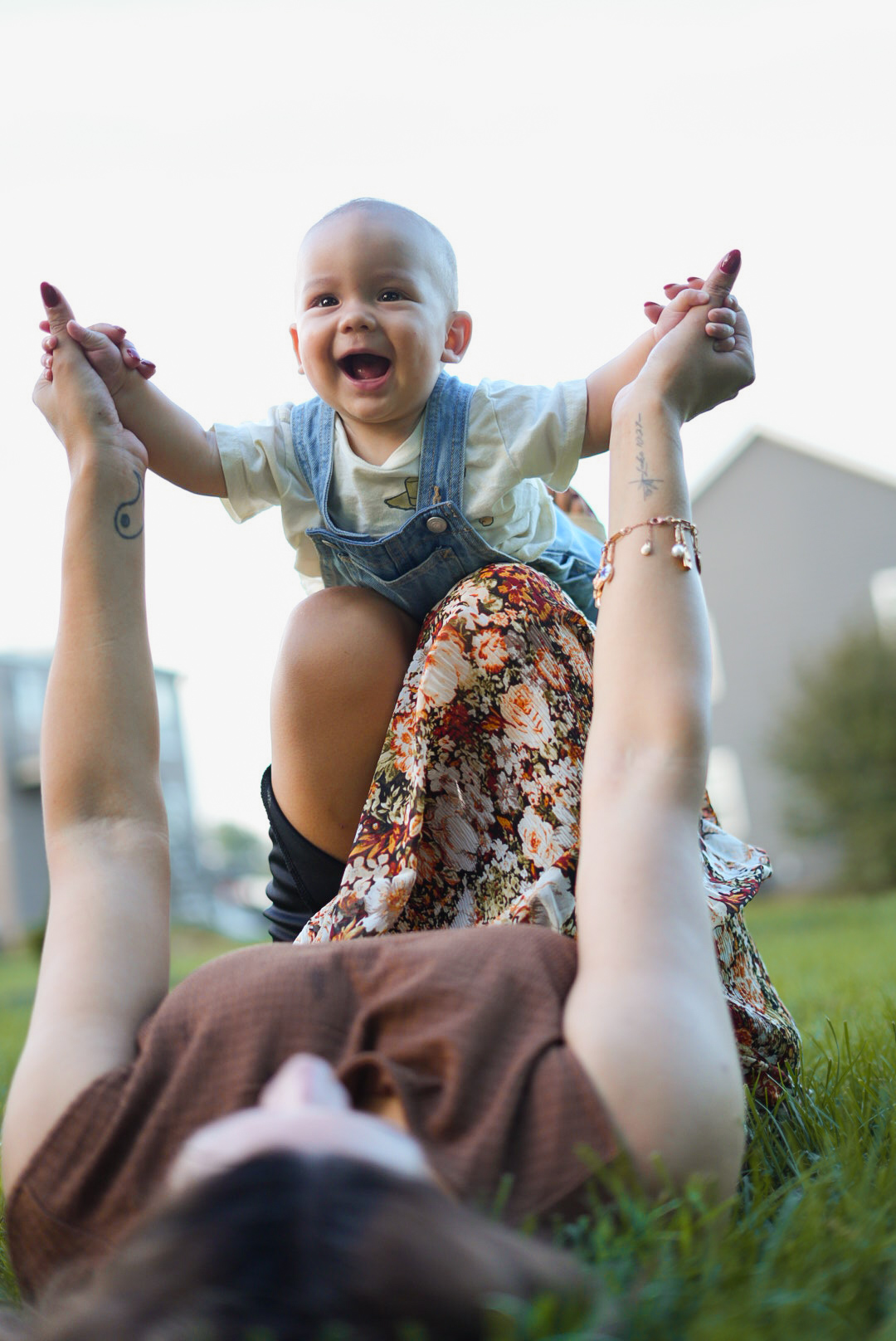 Child with arms raised, smiling, held by an adult lying on grass outdoors, with a building and trees in background.