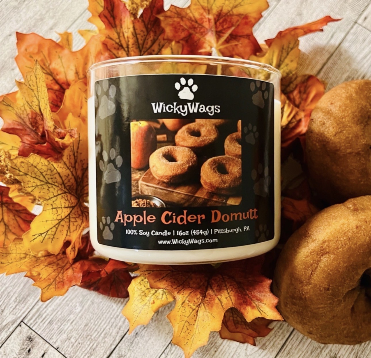 A candle labeled 'WickyWags Apple Cider Donut' surrounded by autumn leaves and potatoes.