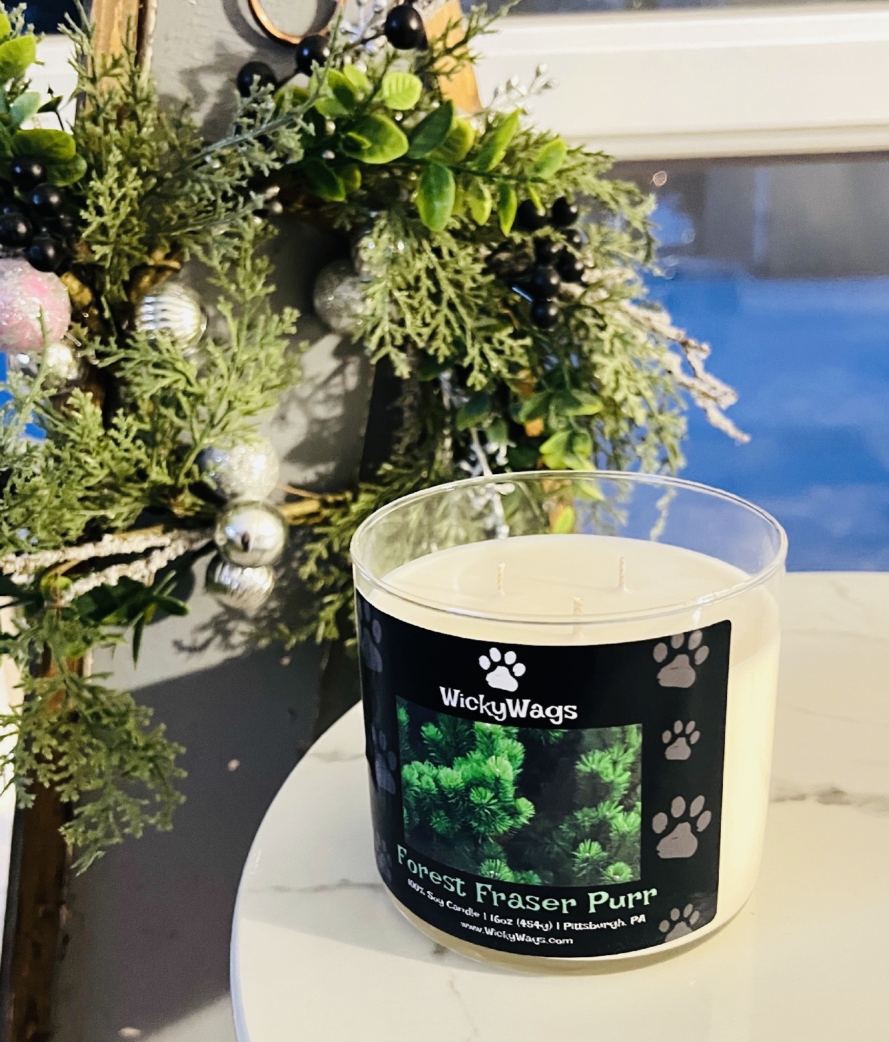 A candle with a label featuring a green dog and paw prints, placed on a white surface near a window with a Christmas decoration nearby.