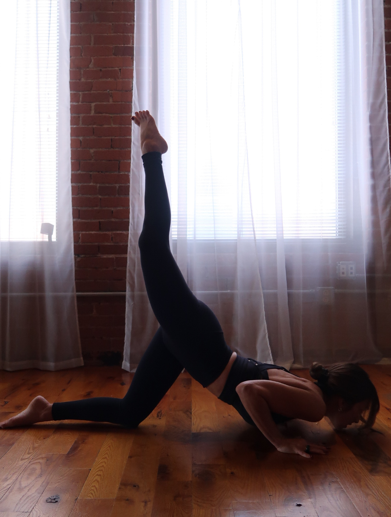 Person performing a yoga pose on the floor with one leg raised vertically, head and hands on the ground, in front of curtains and brick wall.