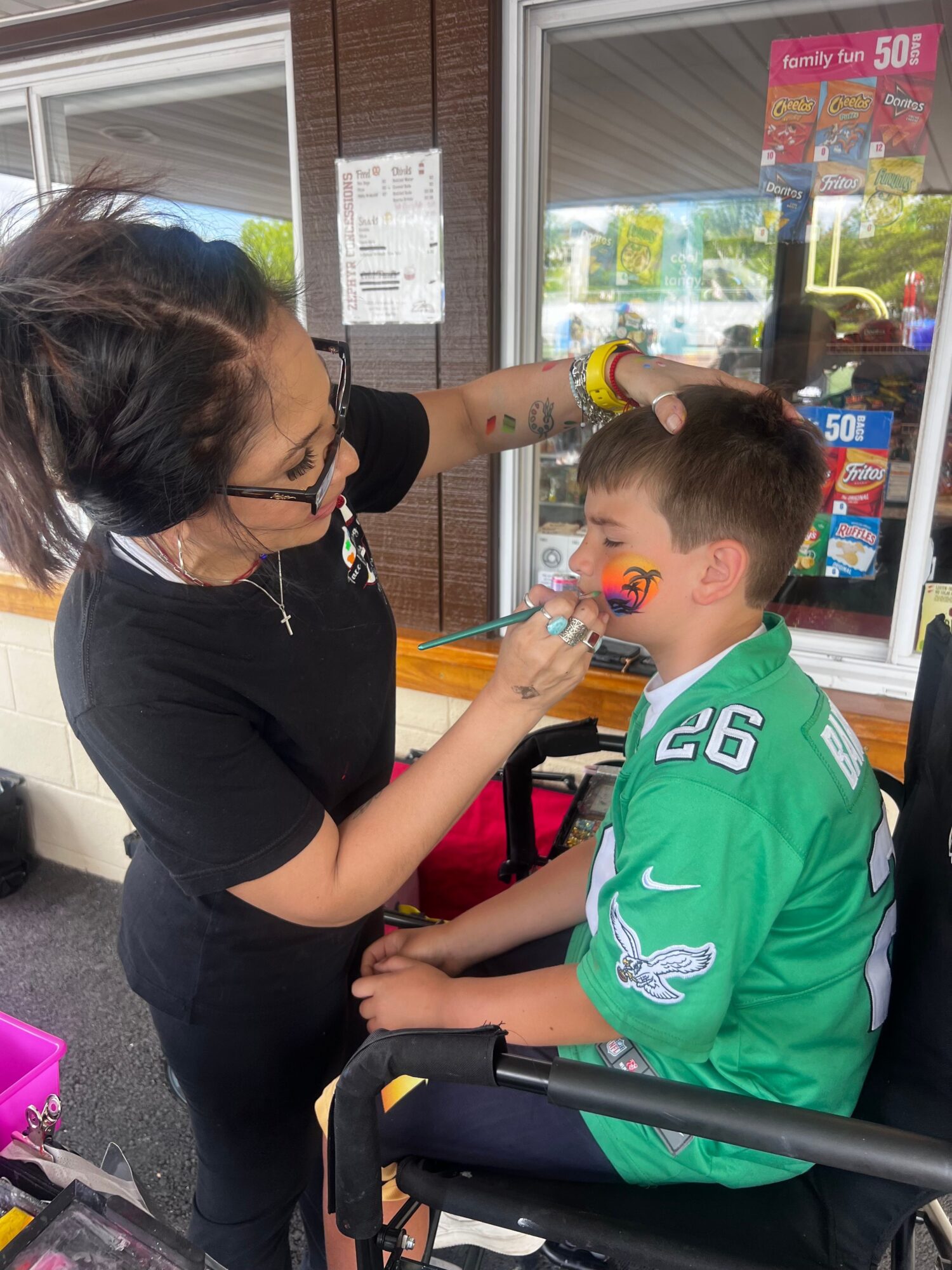 A woman with glasses and a face mask is painting a child's face at a store counter, with shelves and a window in the background.
