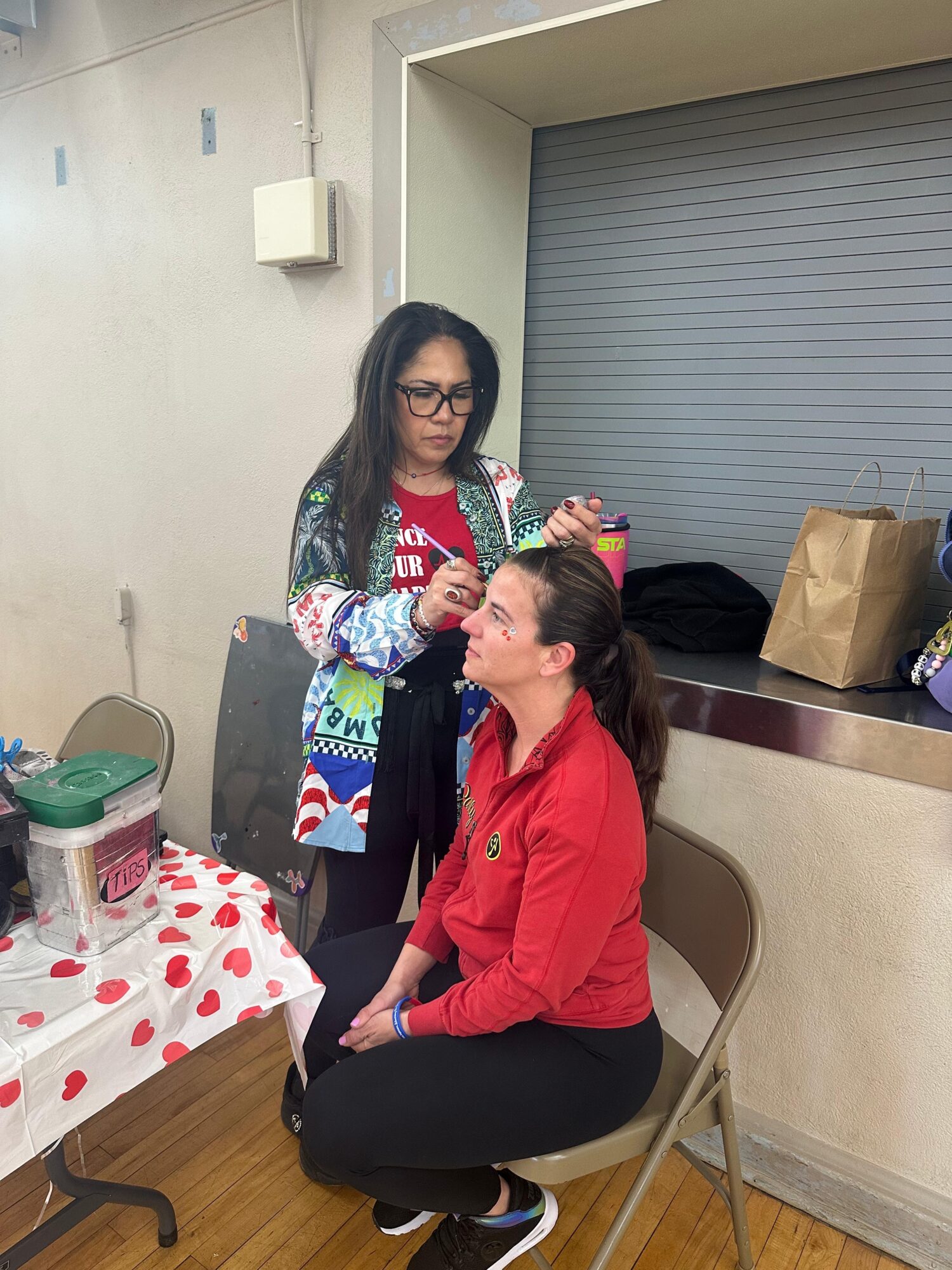 Two women, one standing and one sitting, in a room with a table and a closed gray shutter door. The standing woman is applying makeup or styling the seated woman's hair. The seated woman wears a red jacket, and the standing woman wears glasses and a colorful sweater. A table with a polka dot tablecloth and various items is visible on the left.