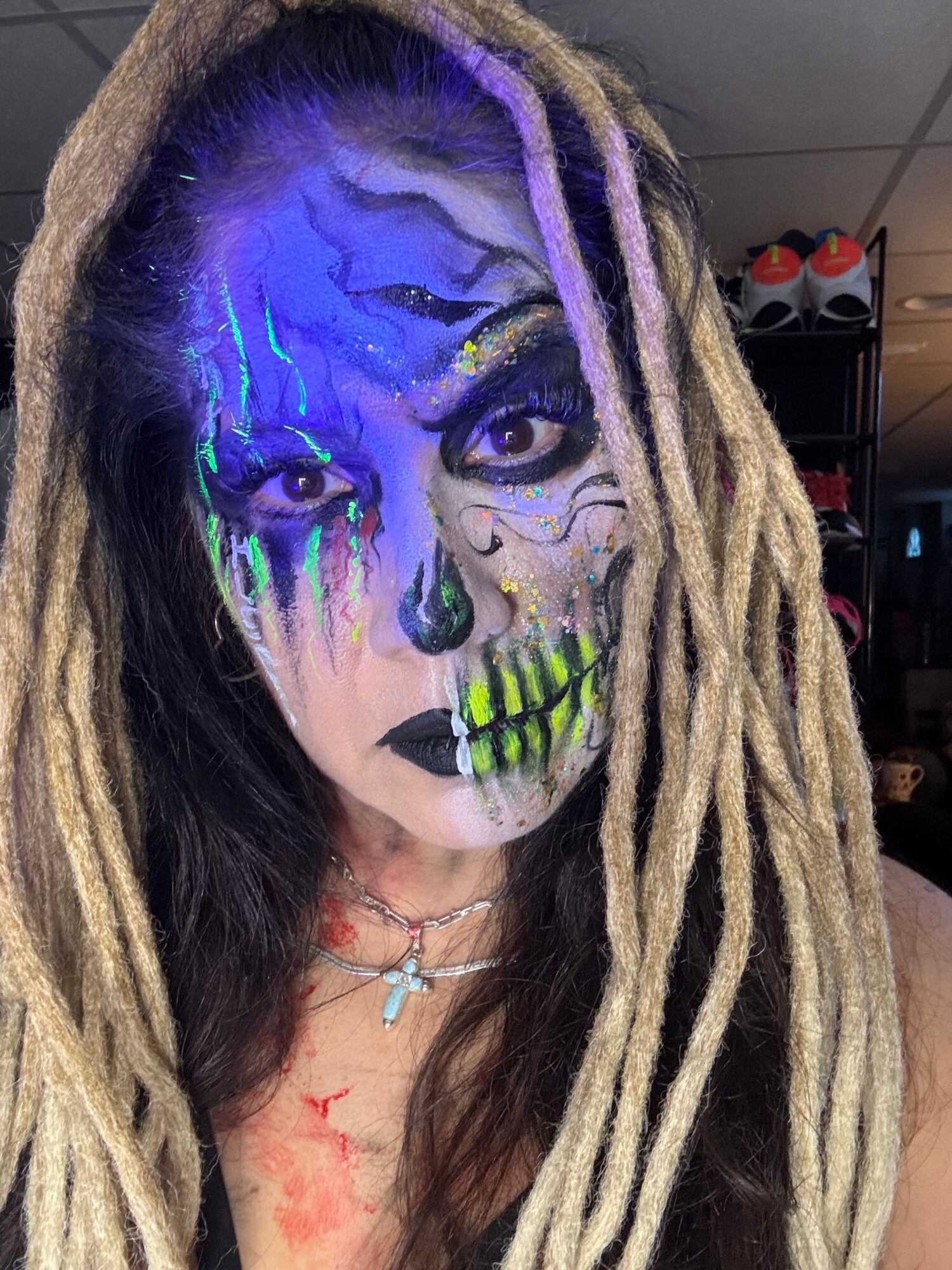 Person with face painted as a skeleton, with long dreadlocks, wearing a cross necklace, in a room with a traffic light in the background.