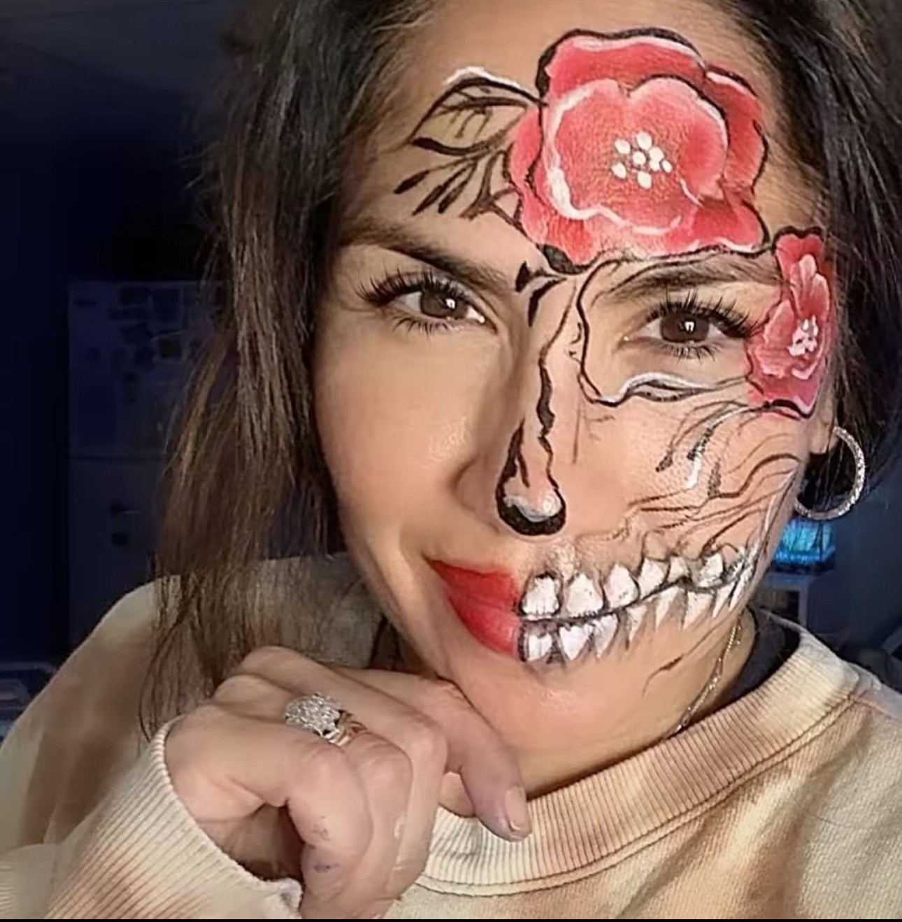 Woman with face painted half as a skull and half with flowers, smiling, hand near chin, wearing a beige top.