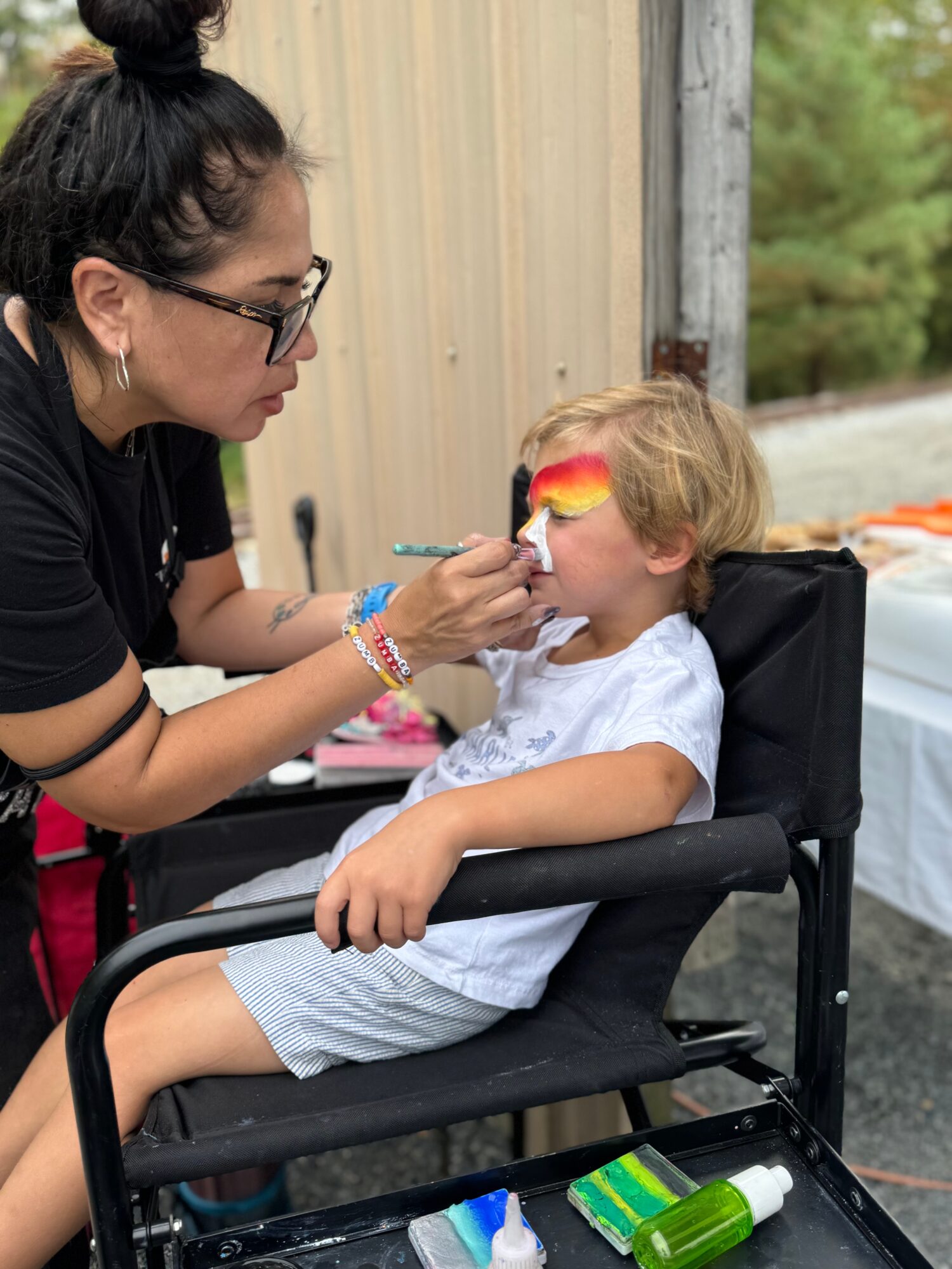 Woman painting a child's face with rainbow colors outdoors, child sitting in a wheelchair, background of trees and a wooden fence.