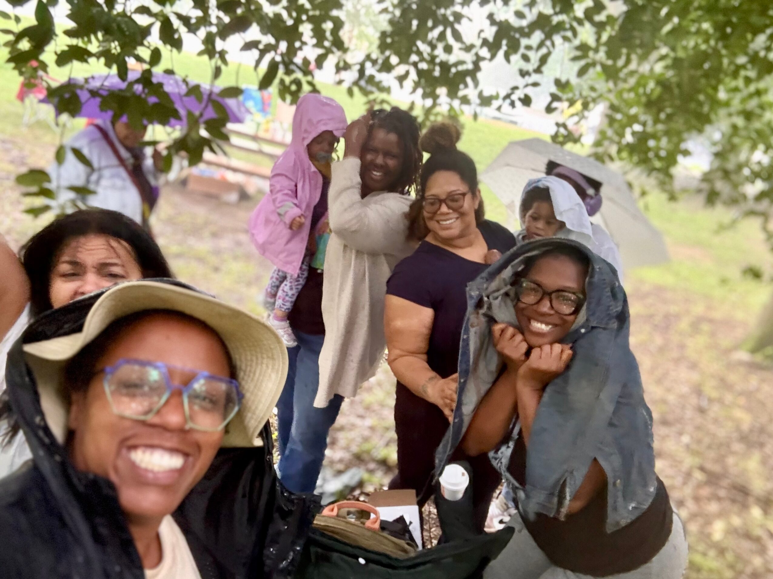 Group of smiling people outdoors, some wearing hats and glasses, under trees with a tent in the background.