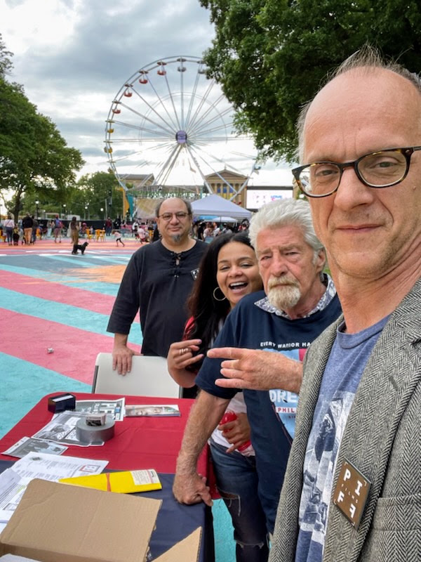 Four people at an outdoor event with a Ferris wheel in the background, standing near a table with papers and boxes, smiling.