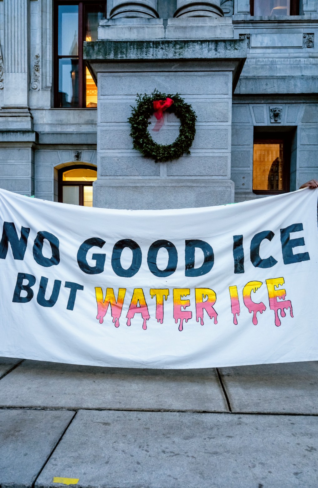 Protest sign with text about ice, water, and climate change, held in front of a building with a wreath.