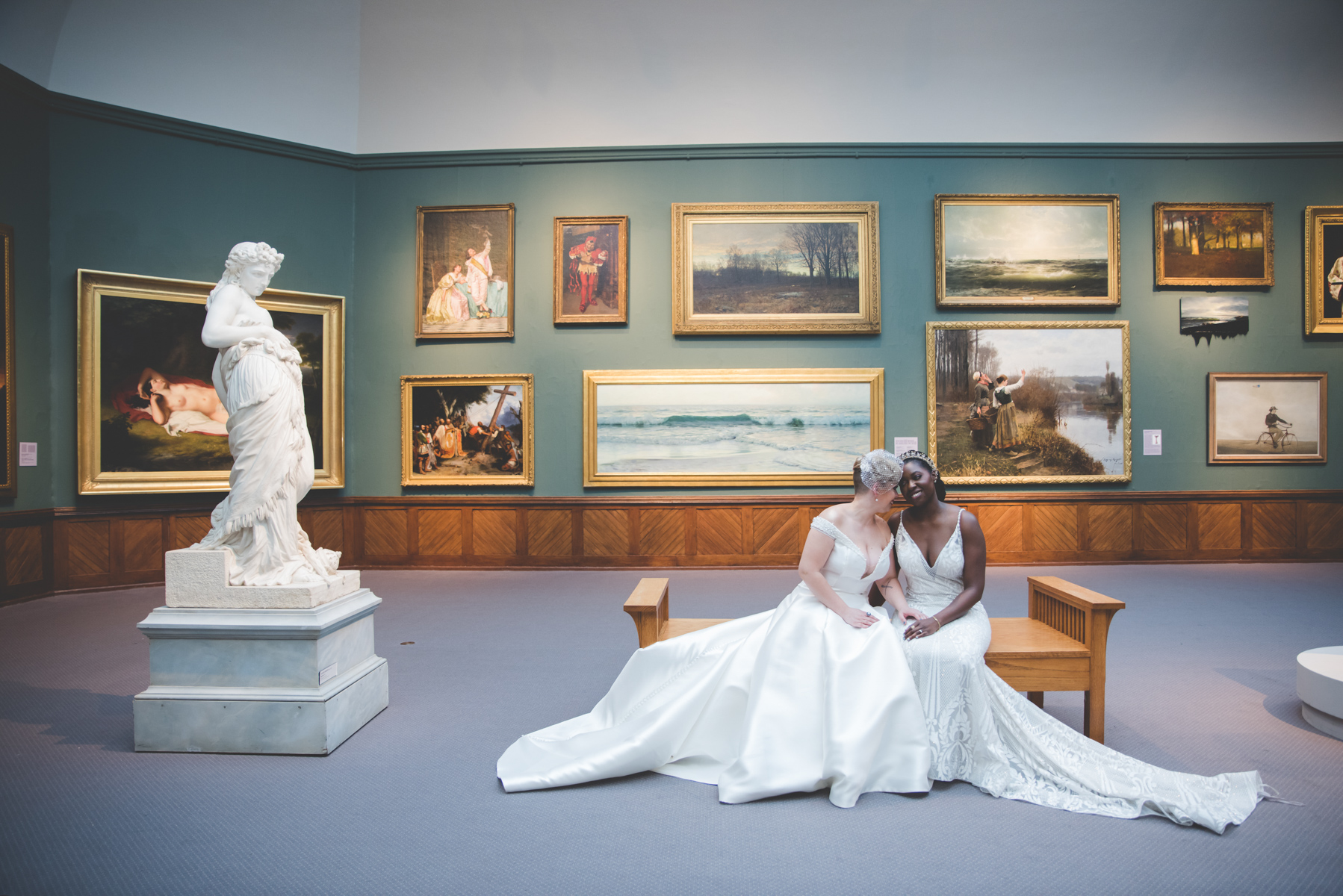 Two women in wedding dresses sit on a bench in an art gallery, surrounded by framed paintings and a sculpture. Alt text word count: 20. Confidence: 0.9