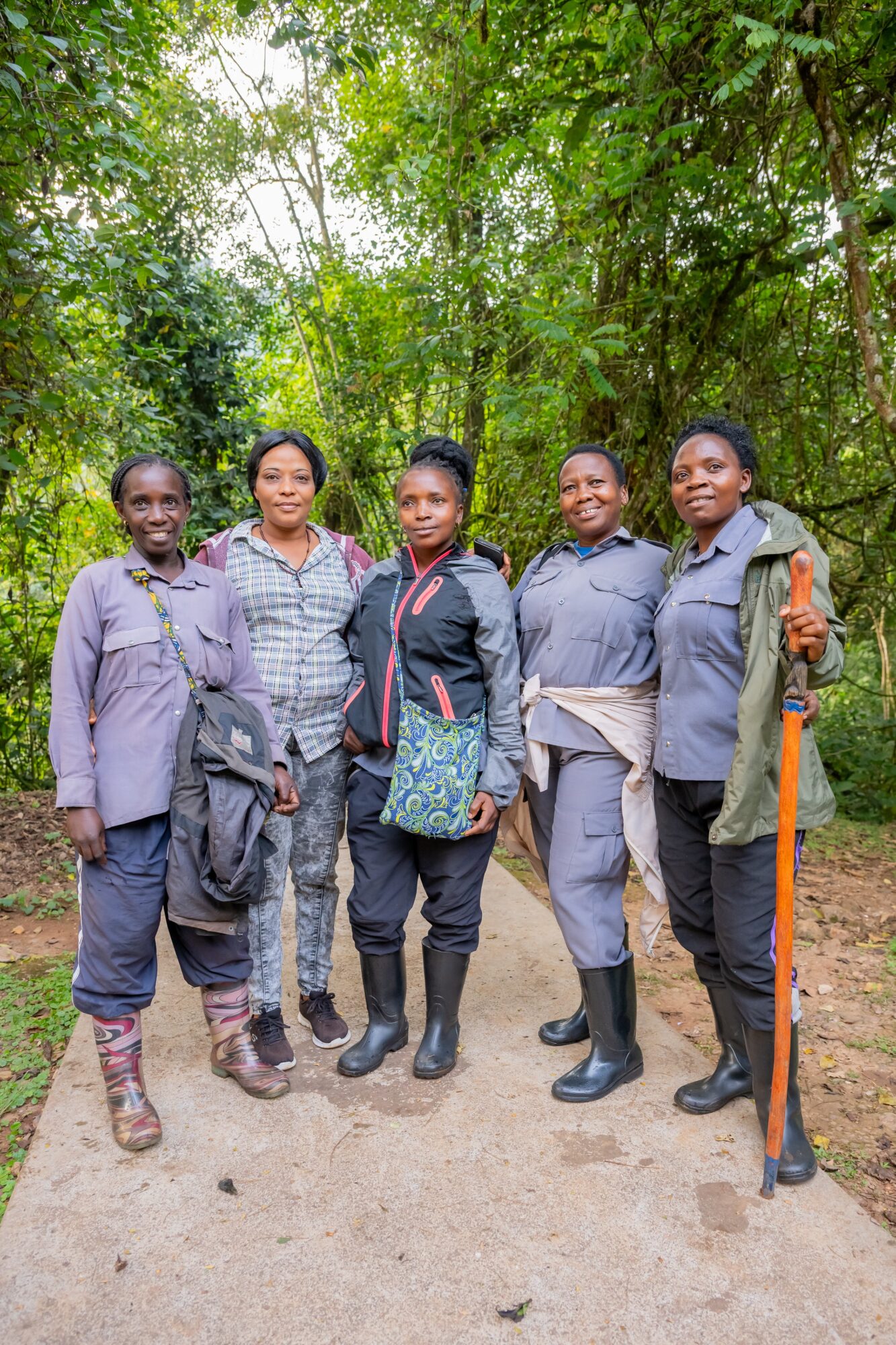 Five women standing outdoors on a dirt path surrounded by green trees, smiling and posing for the photo.