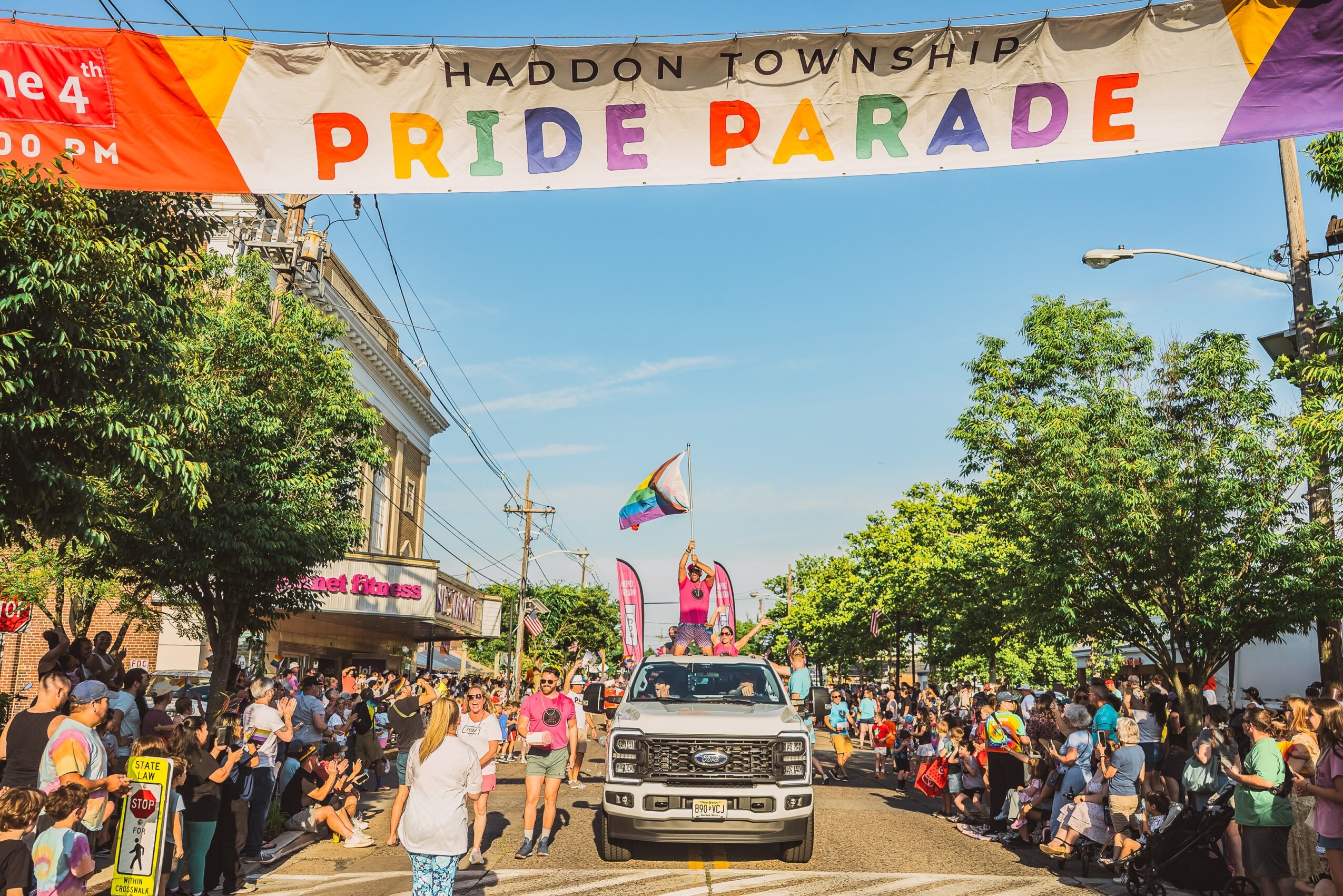 Crowd gathered along a street during Pride Parade with a colorful banner overhead, trees, buildings, and a vehicle in the street.