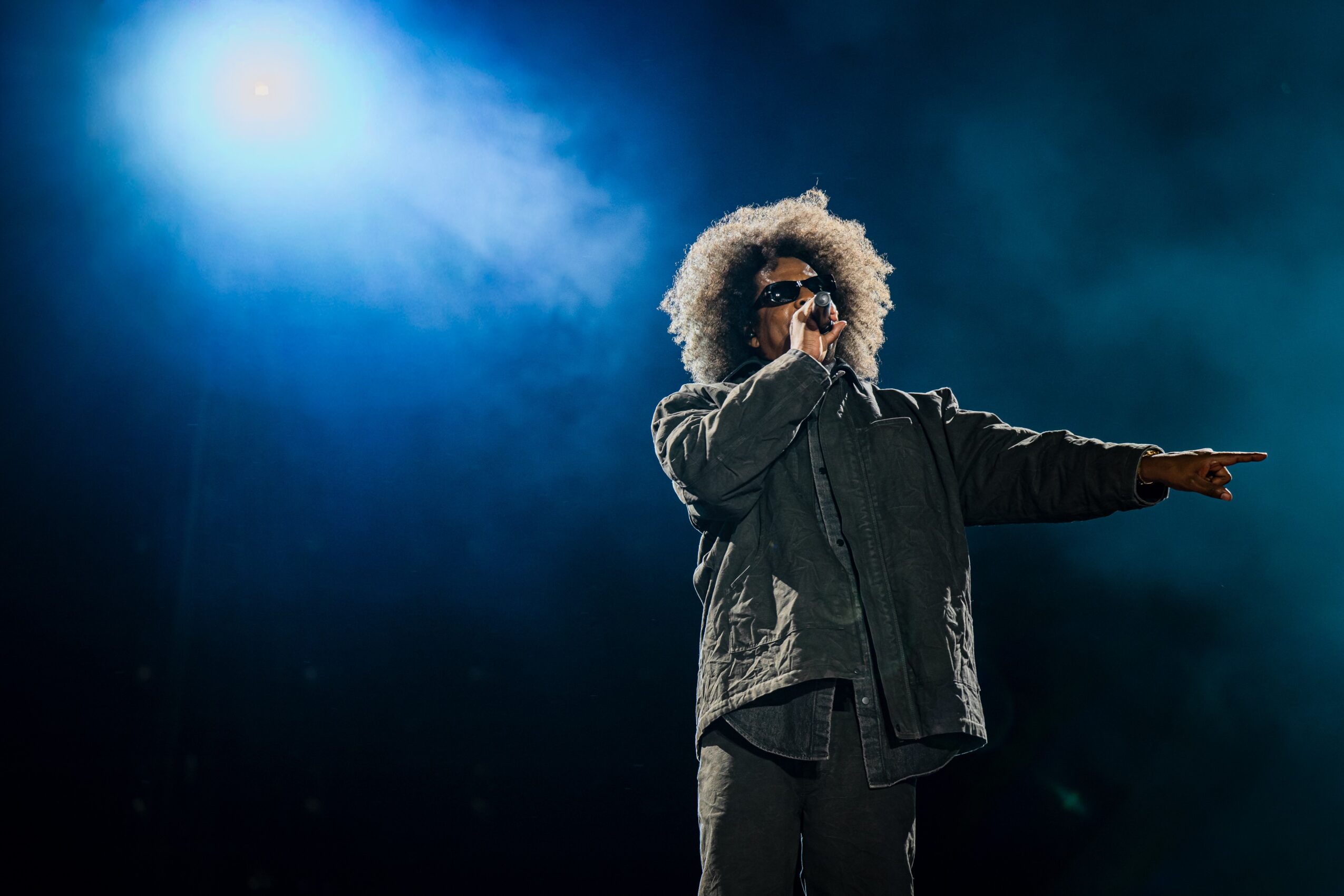 Person with curly hair and sunglasses performs on stage with a dark background and bright light, holding a microphone.