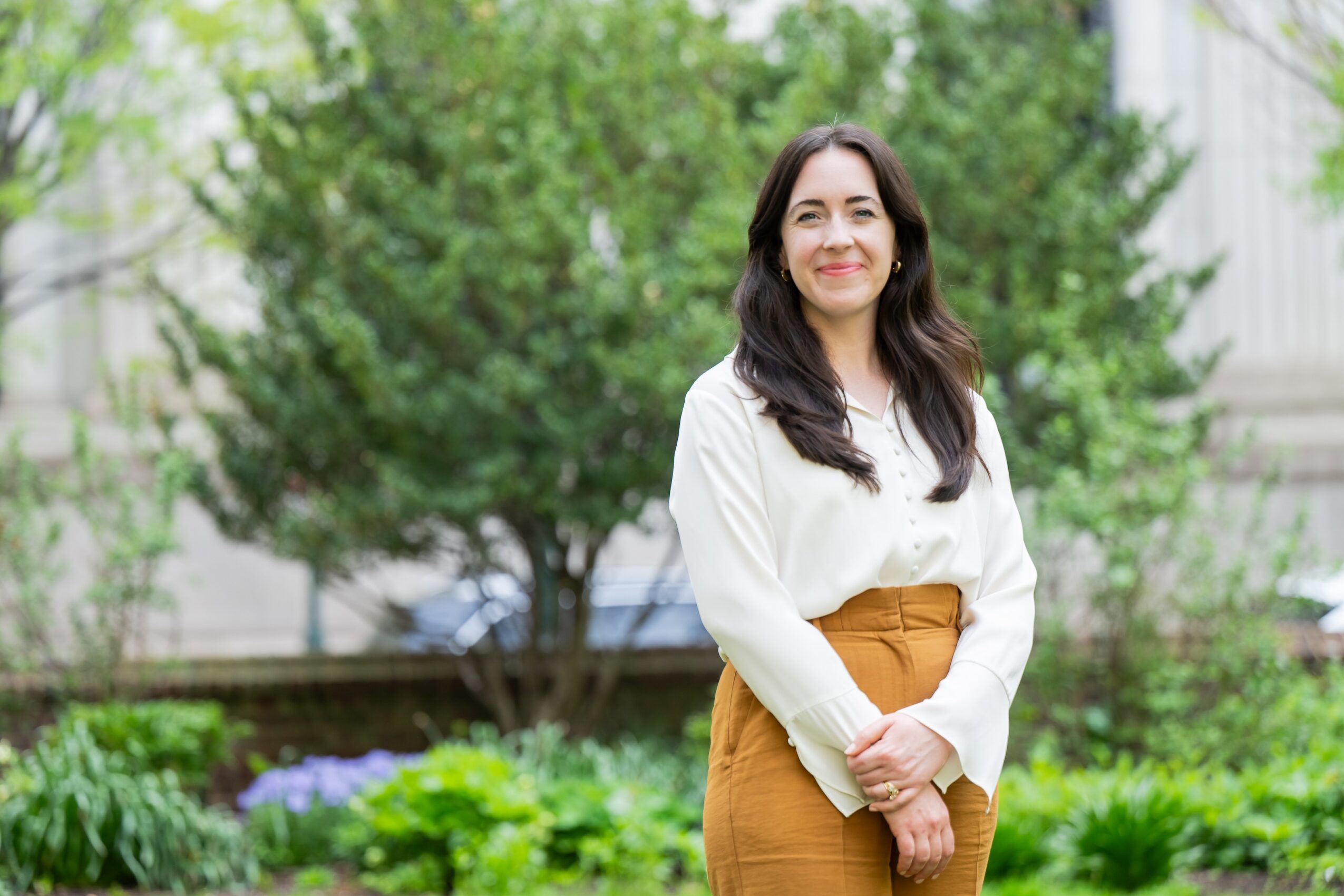Woman standing outdoors in a garden with green trees and plants, smiling, wearing a white blouse and brown skirt.