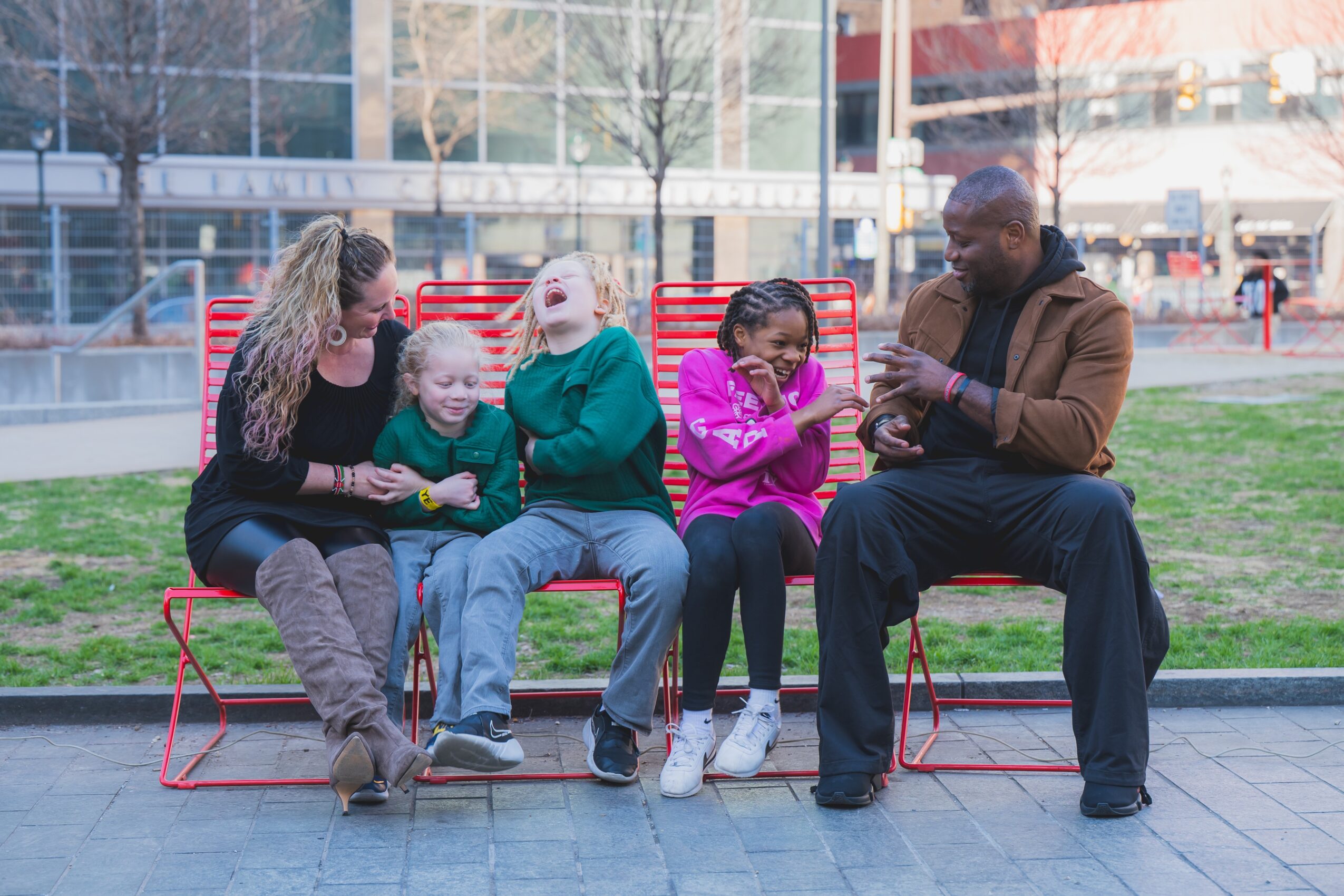 Group of five people sitting on red benches outdoors, engaging with their phones and talking, with a cityscape background.
