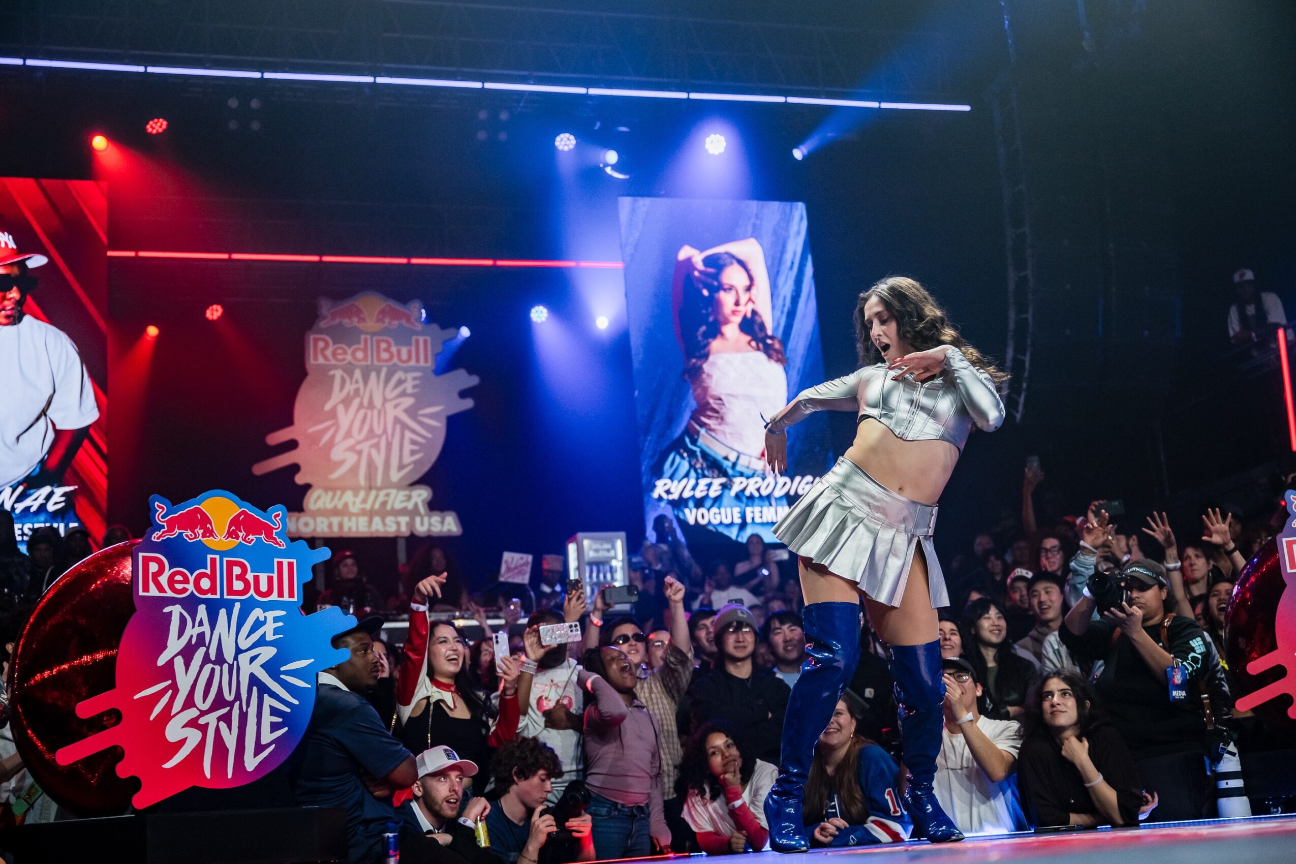 Two performers dance on stage with a large crowd watching, colorful lights, and promotional banners in the background.
