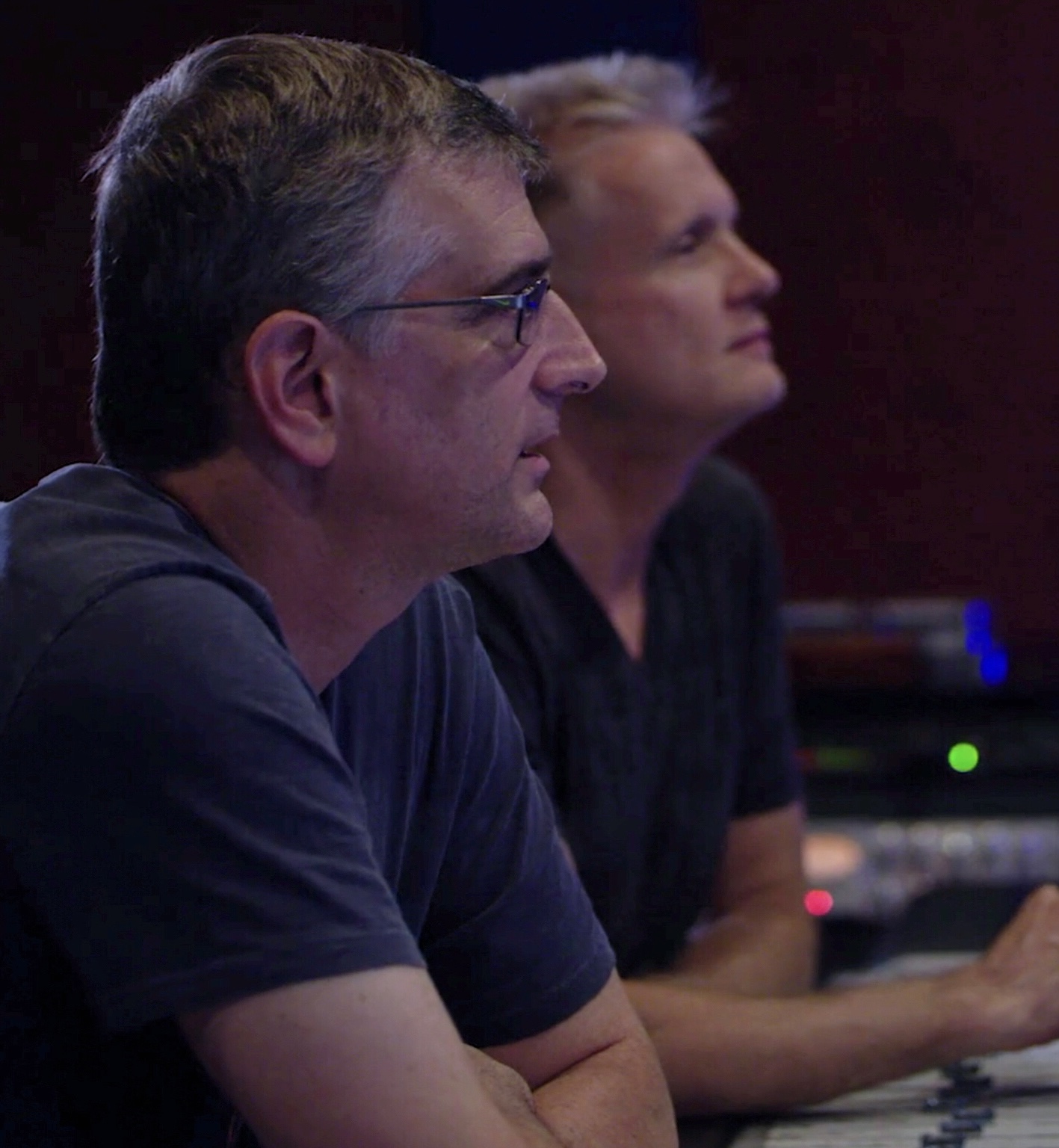 Two men sitting side by side, focused, in a dimly lit room with equipment in the background.