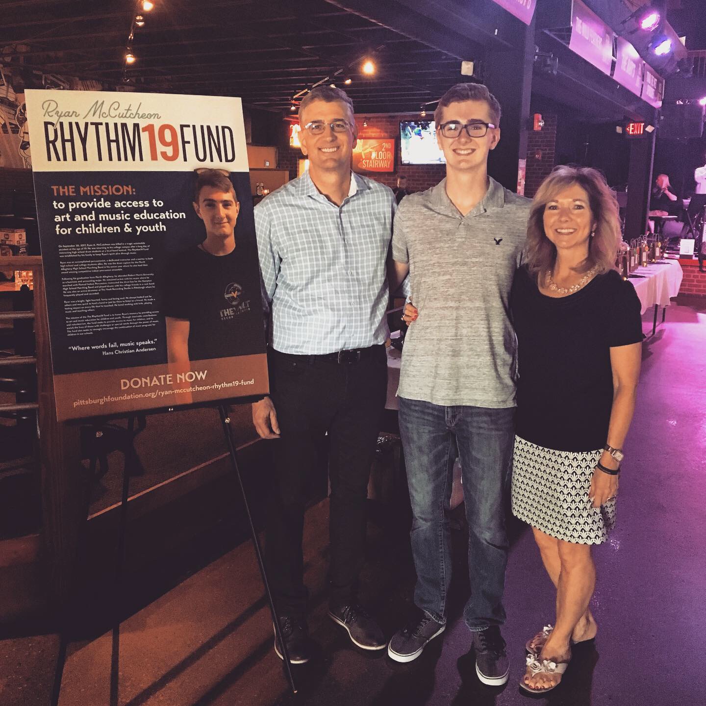 Three people standing together indoors, smiling, with a sign about a music education fund in the background.