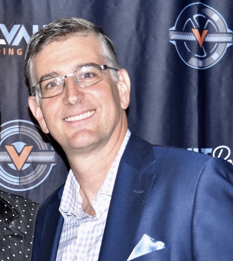 Man with glasses smiling, wearing a suit and tie, standing in front of a dark backdrop with logos.
