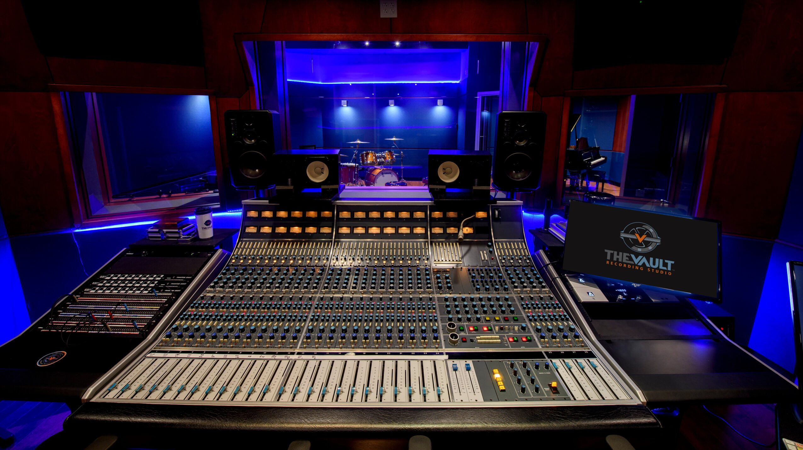 Recording studio mixing console with speakers and a view of a stage through windows, illuminated by blue lighting.