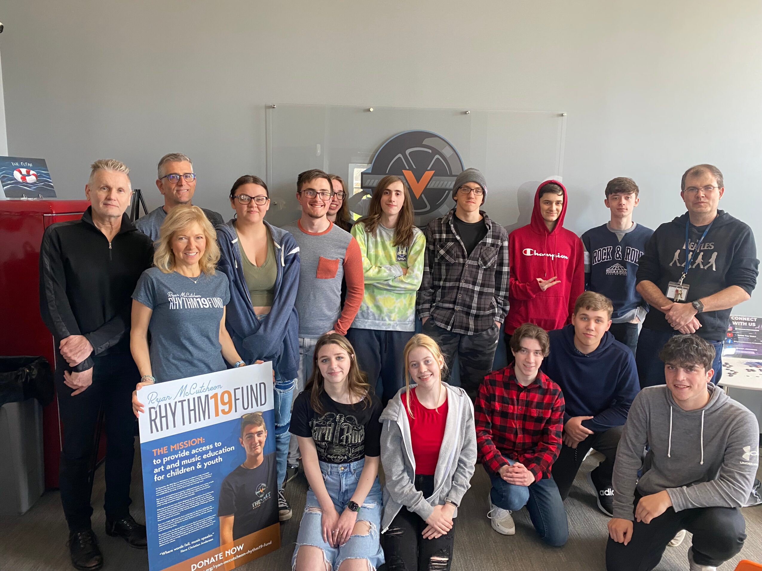 Group of fifteen people standing indoors, some holding a sign, with a logo on the wall behind them.