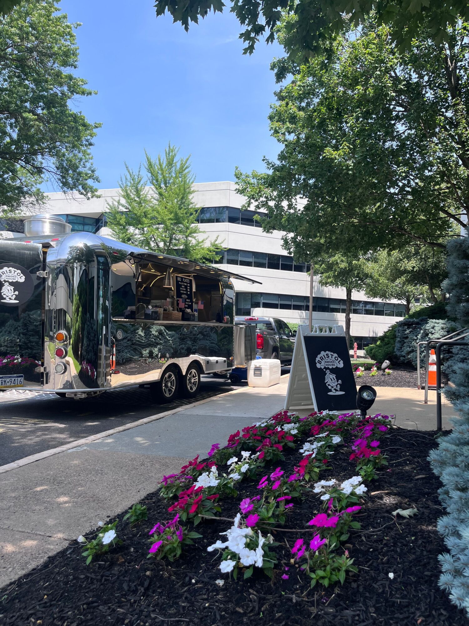 A food truck parked on a street with trees and flowers nearby, under a clear blue sky.