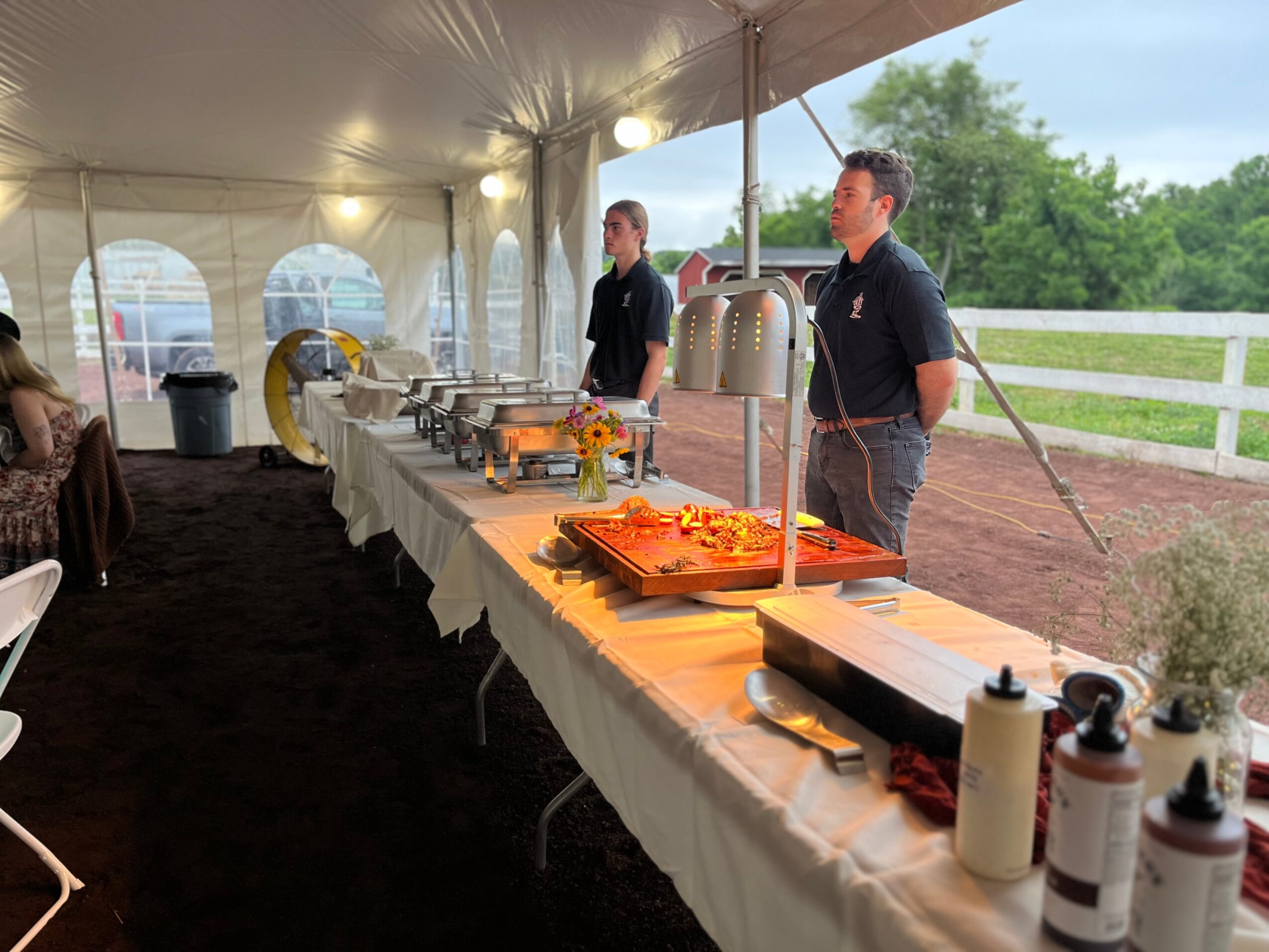 Two men stand behind a buffet table under a tent, with food trays and condiments, outdoors with trees and a fence.