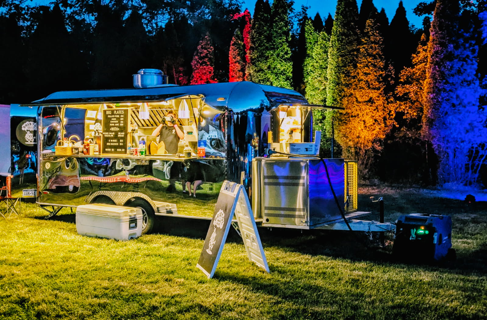 Food truck parked on grass with colorful trees in background, illuminated at night, with a cooler and sign outside.