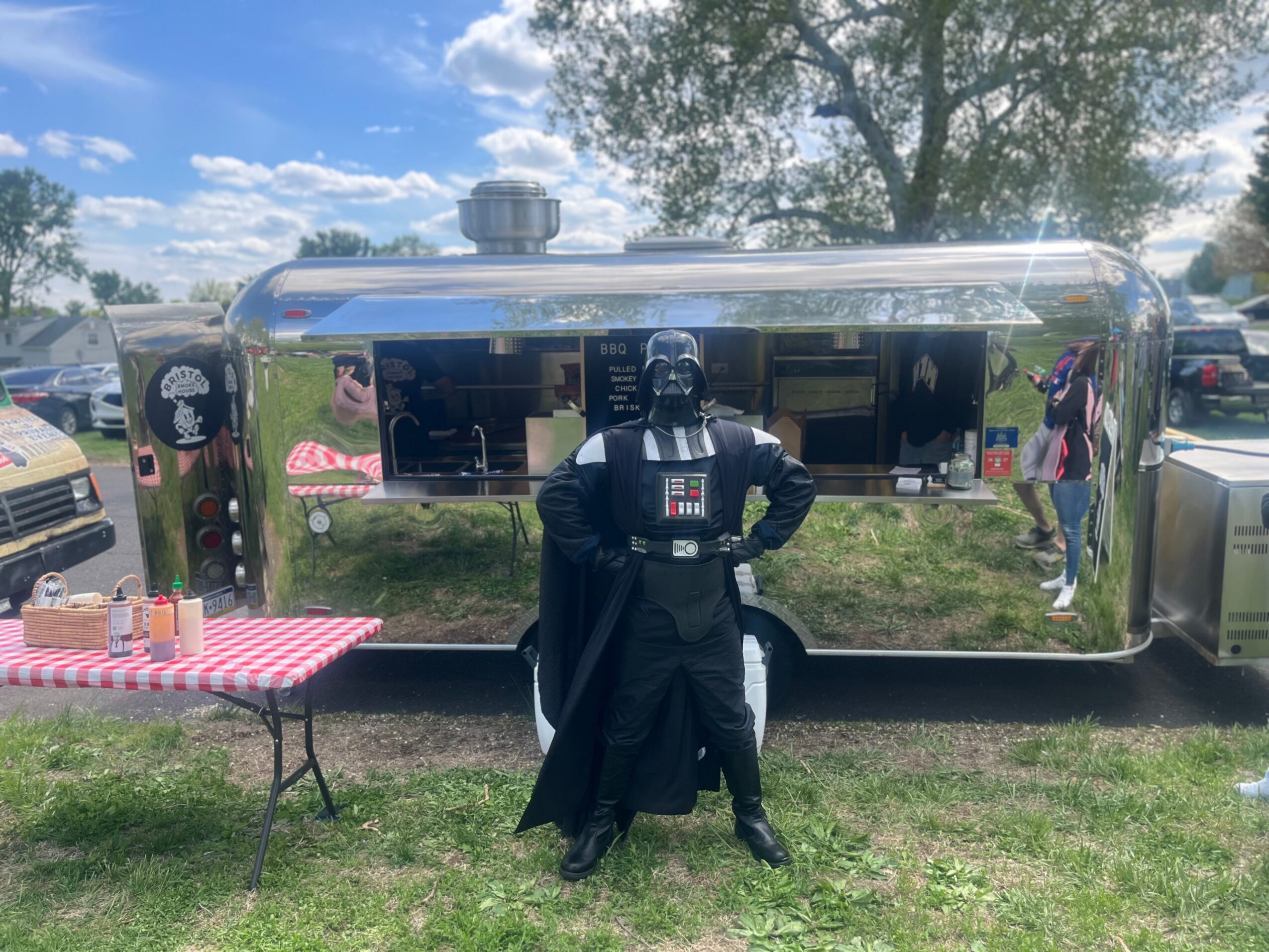 Person dressed as Darth Vader stands in front of a shiny food truck with a window, outdoors on grass with trees and cars nearby.