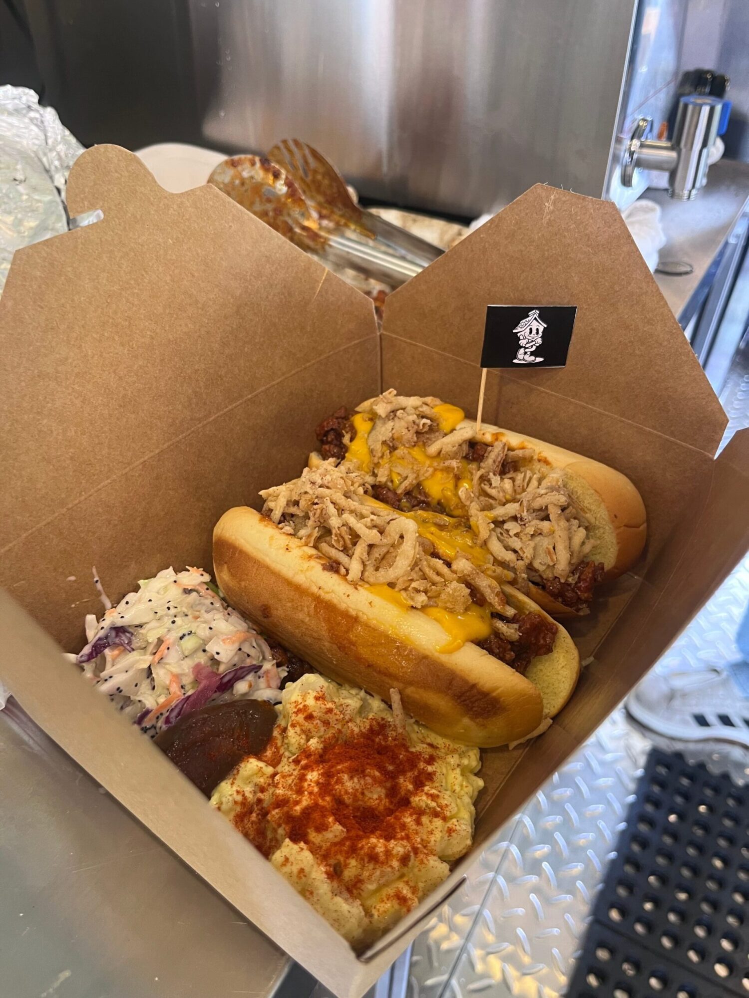 Three hot dogs with toppings in a cardboard box on a metal surface, with a black container in the background.