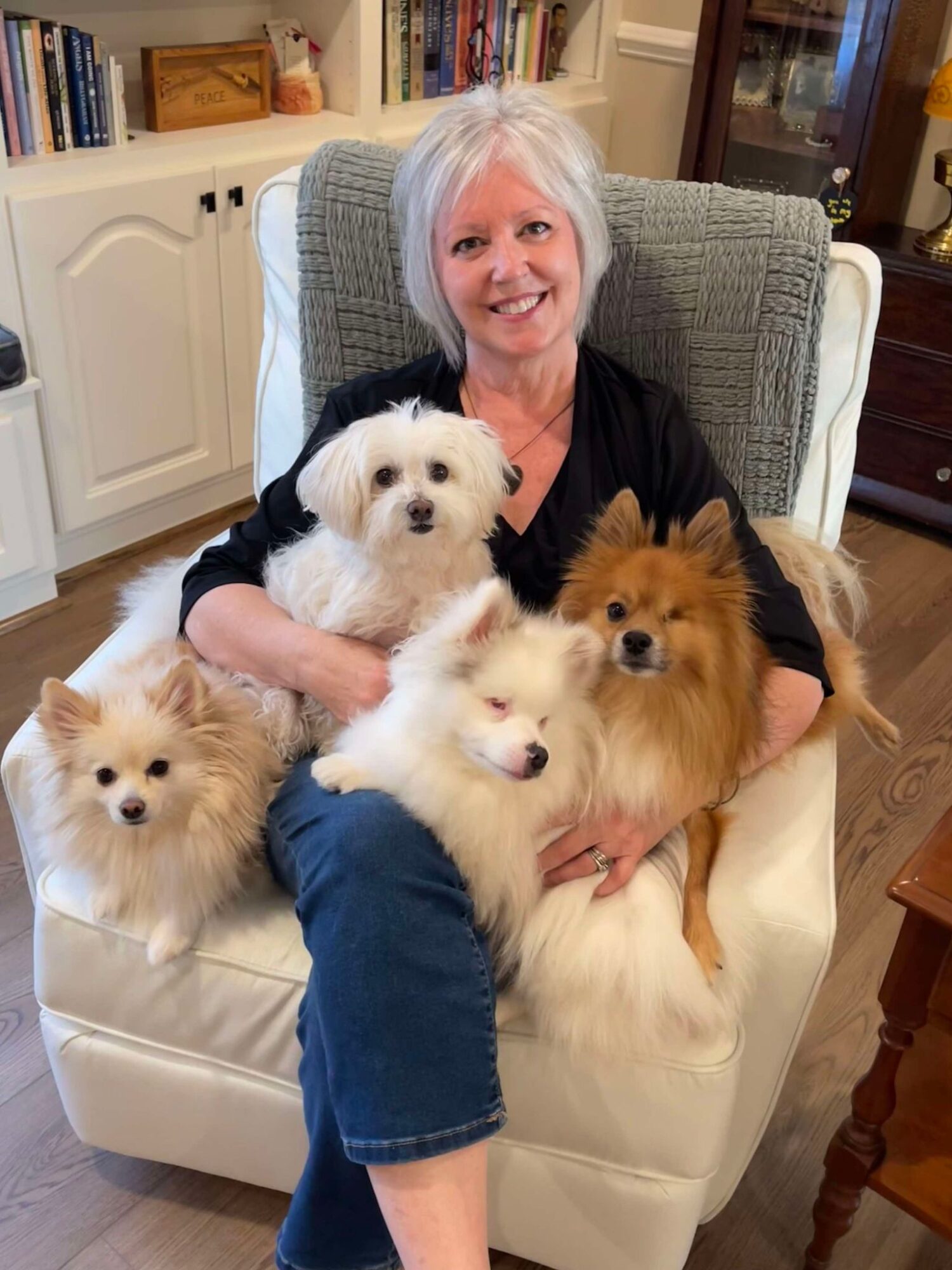 Older woman with white hair sitting in a chair, surrounded by five small dogs, in a cozy room with bookshelves.