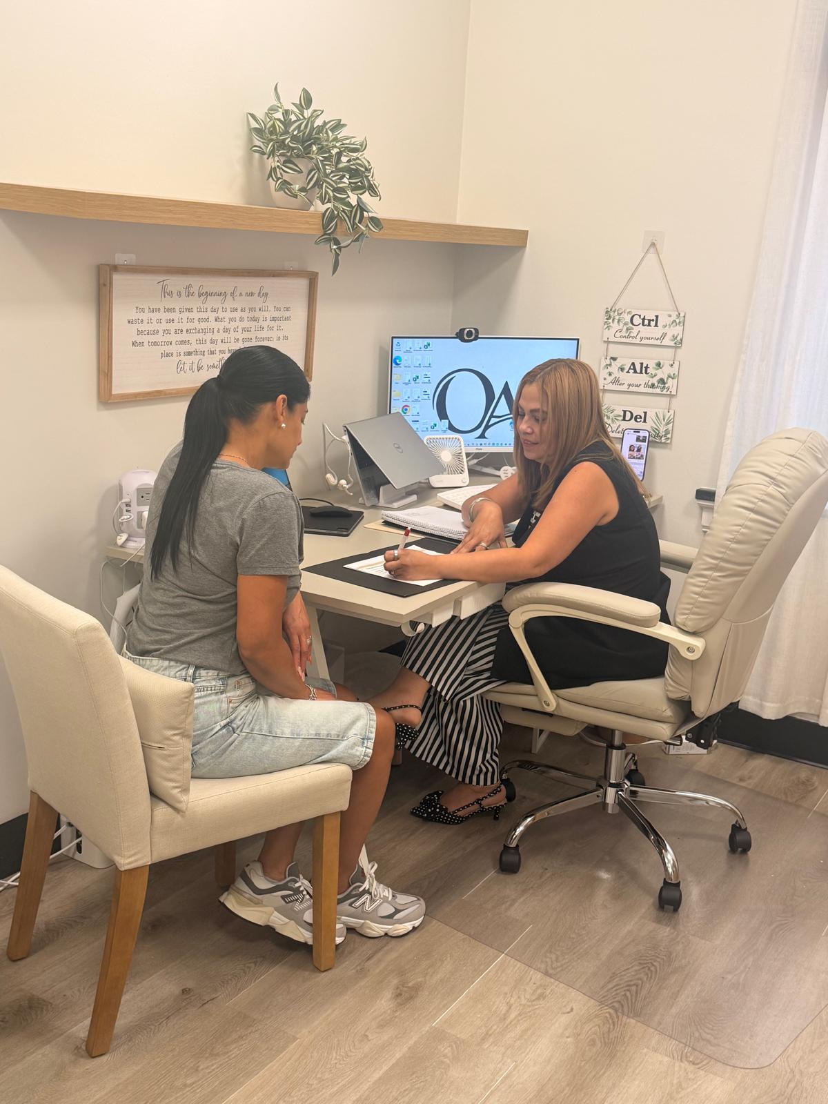 Two women sit at a desk in an office, one working on a computer, the other listening. Office decor includes a plant and framed art.