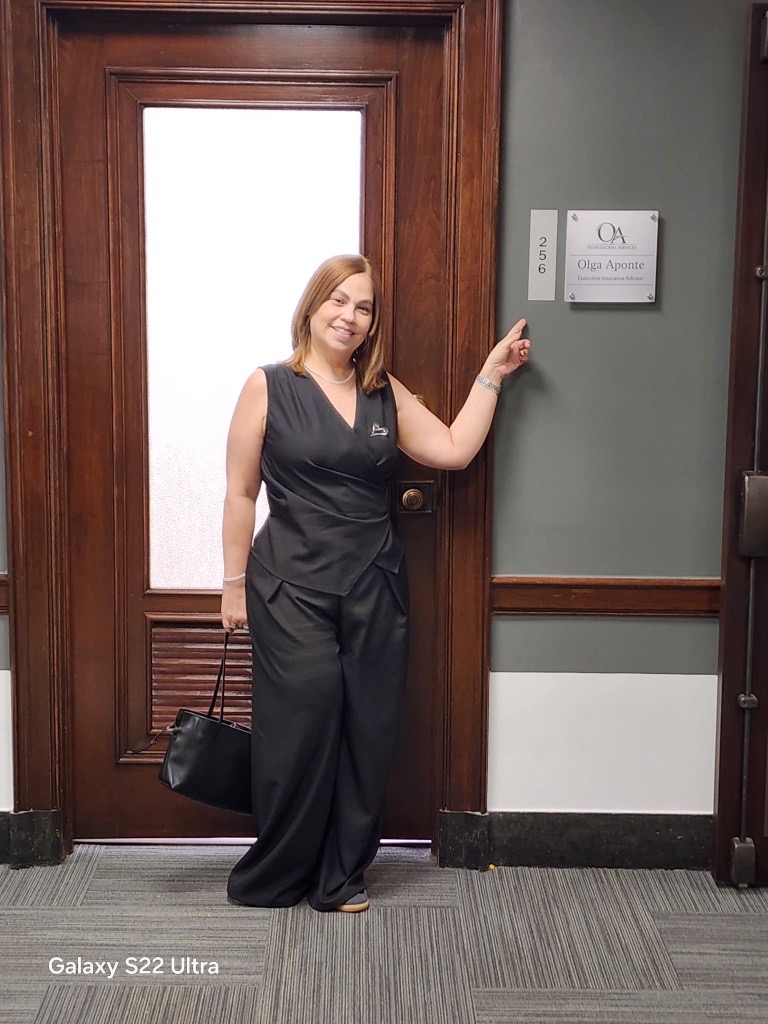 Woman in black outfit standing next to a door, pointing at a sign on the wall, holding a black bag, smiling.