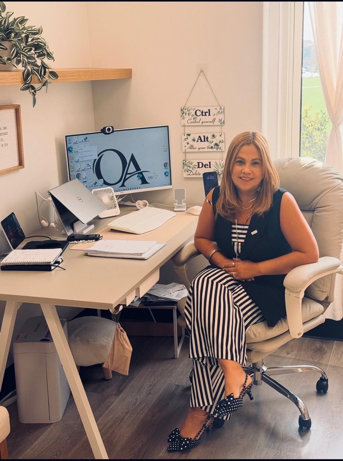 Woman sitting in an office chair at a desk with a computer, books, and office supplies, in a bright room with a window.