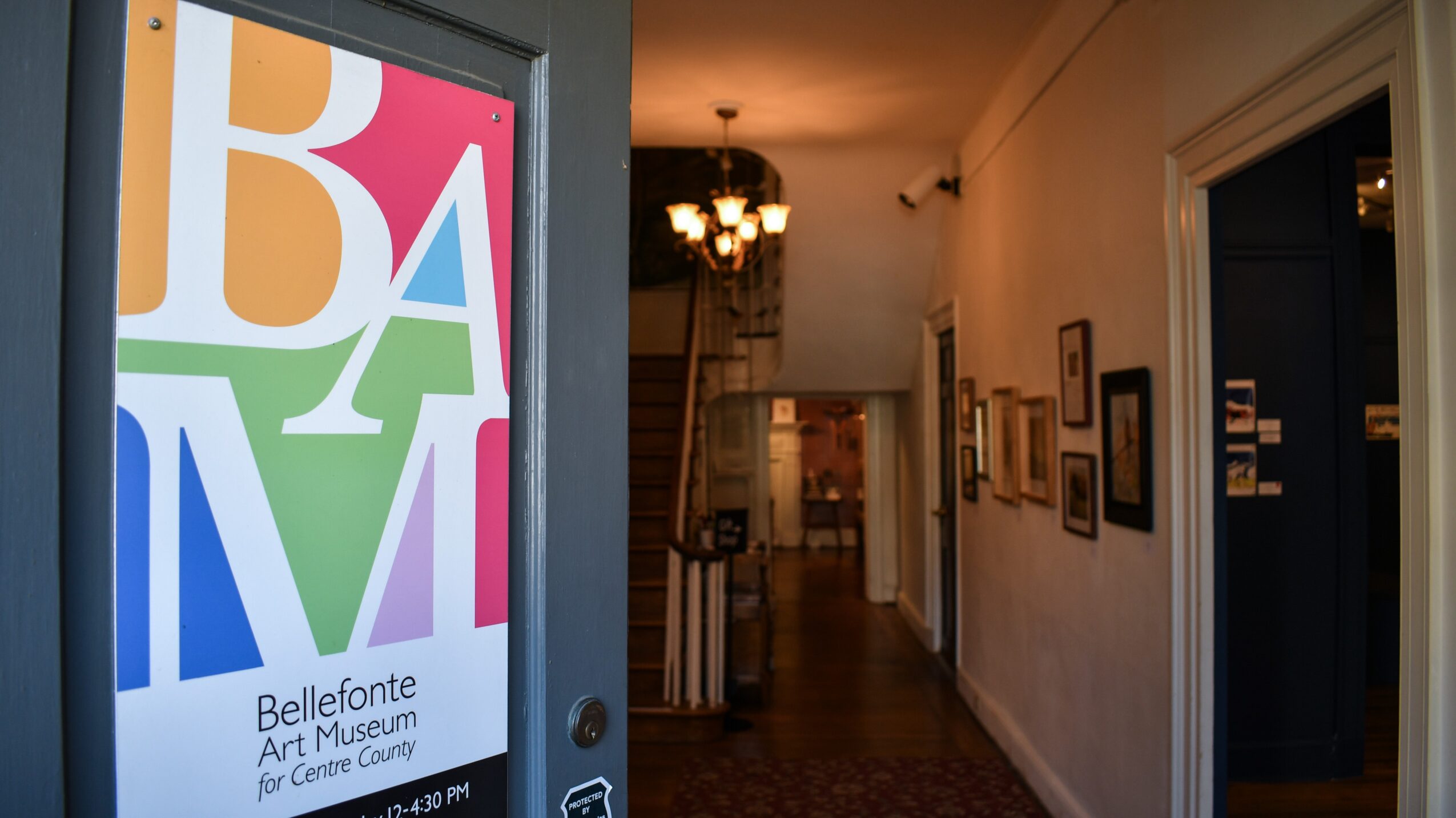 Interior hallway of an art museum with framed artworks on the wall and a chandelier hanging from the ceiling.