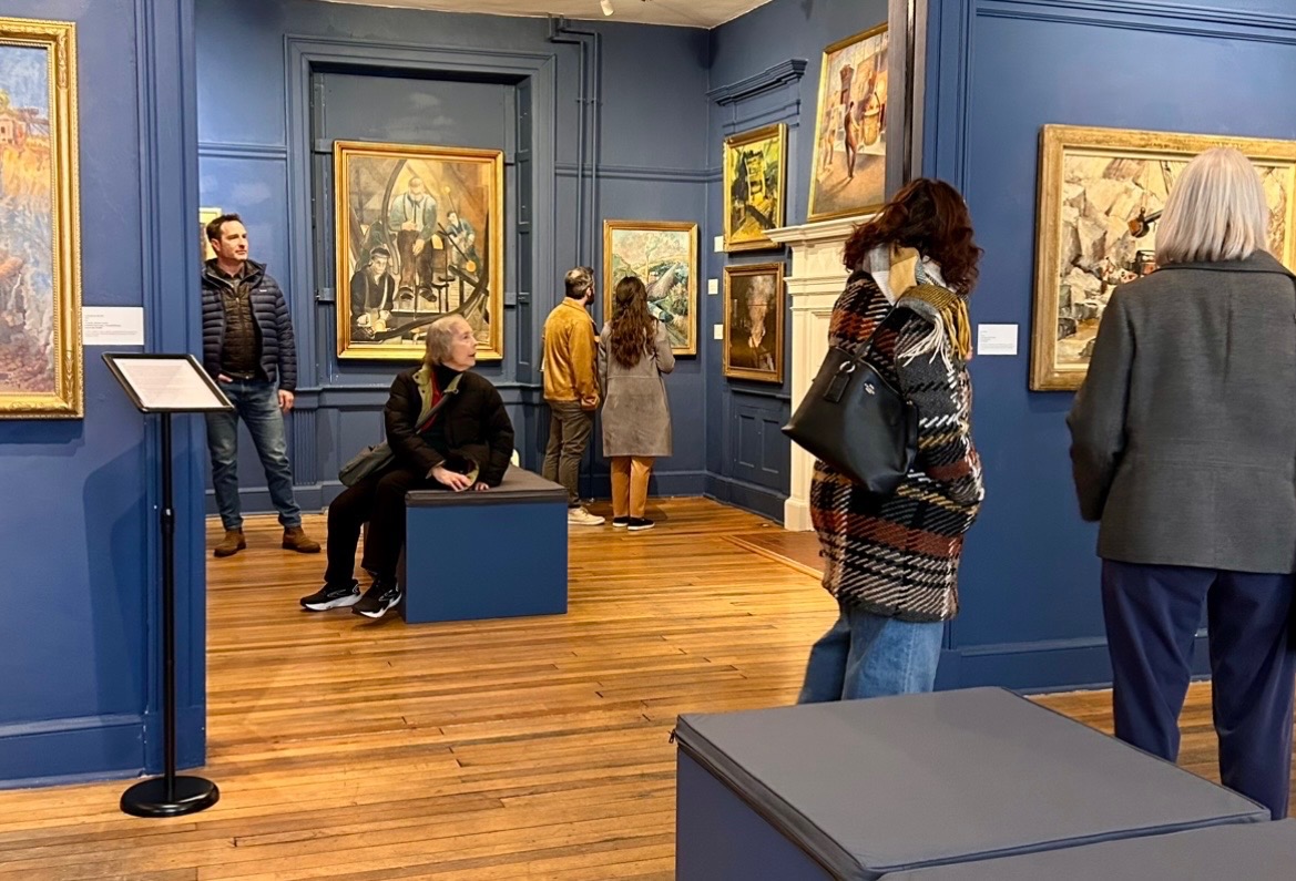 People viewing paintings in an art gallery with blue walls and wooden floors.