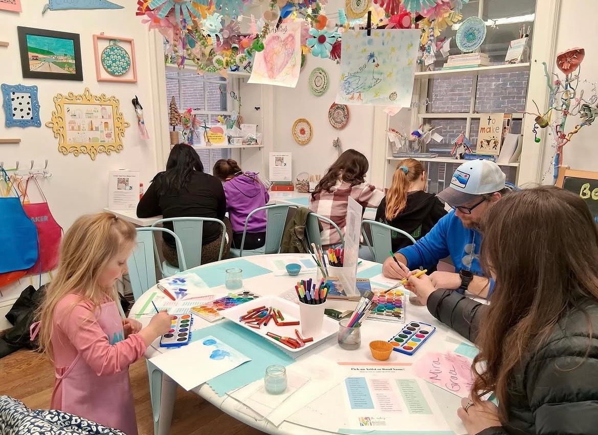 Children and adults sit around a table engaging in arts and crafts, with colorful decorations hanging above in a classroom.