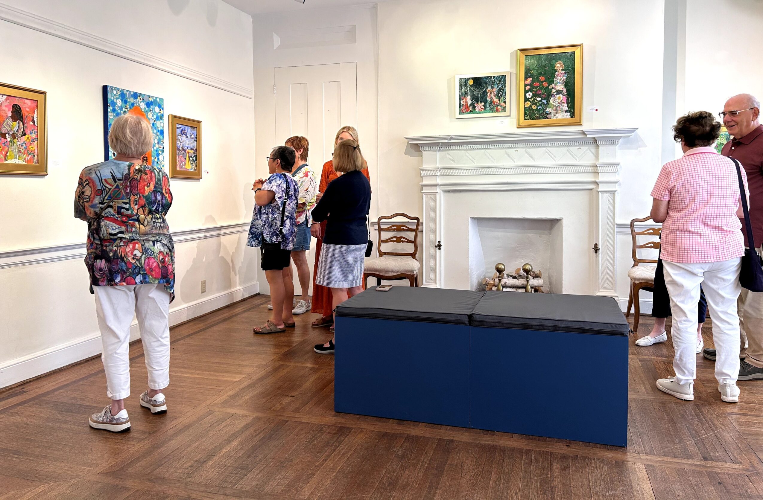 People viewing artwork in an art gallery with paintings on white walls and a fireplace, wooden floor, and a blue bench.