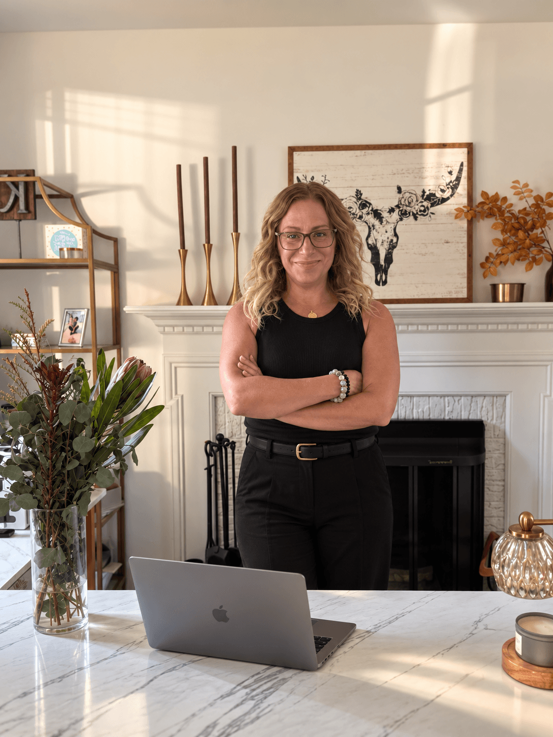 Woman with glasses and curly hair standing with arms crossed in a living room, smiling, with a laptop on a table.
