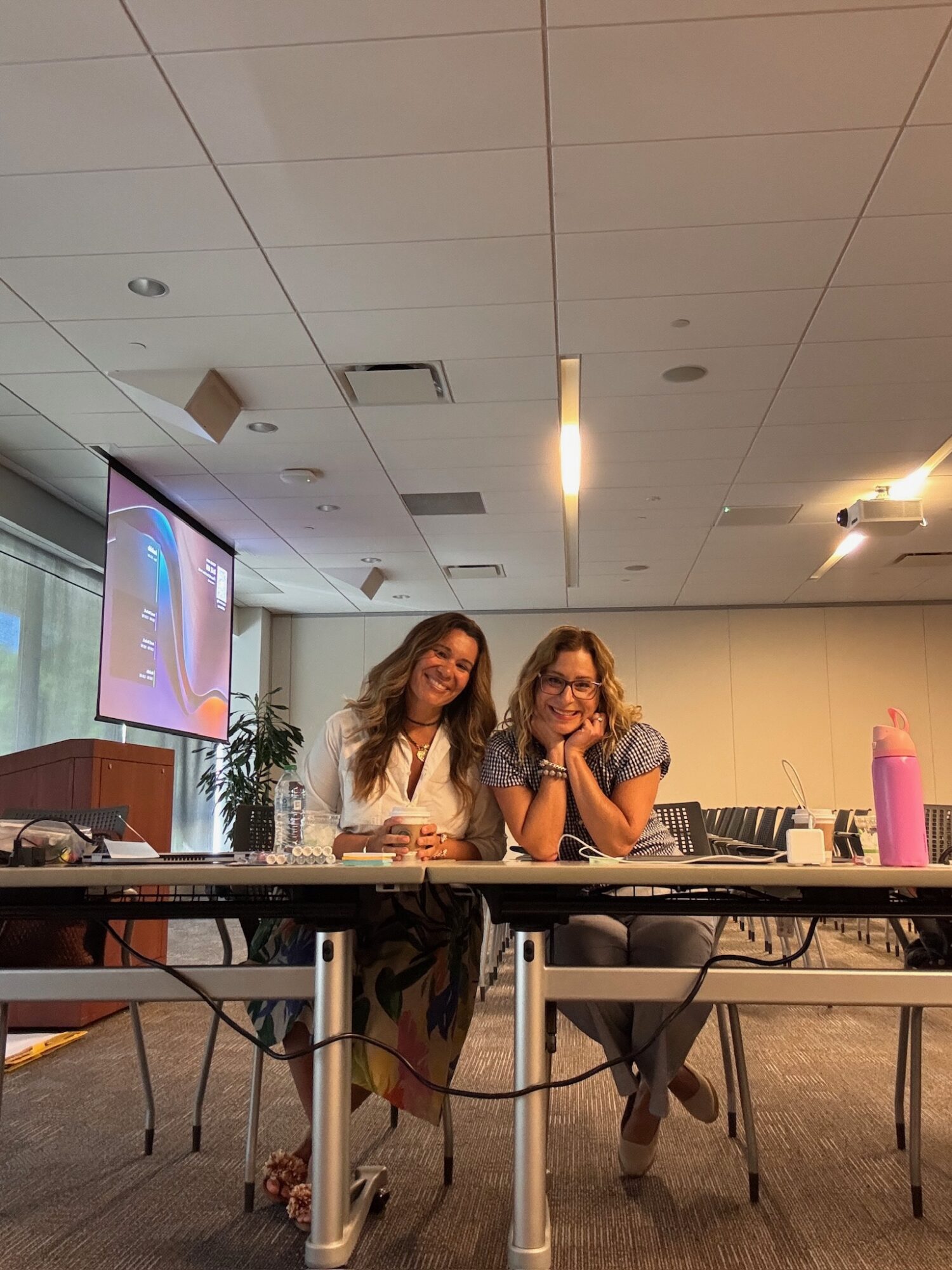 Two women sit at a table in a conference room, smiling and leaning on their hands, with a projector screen behind them.