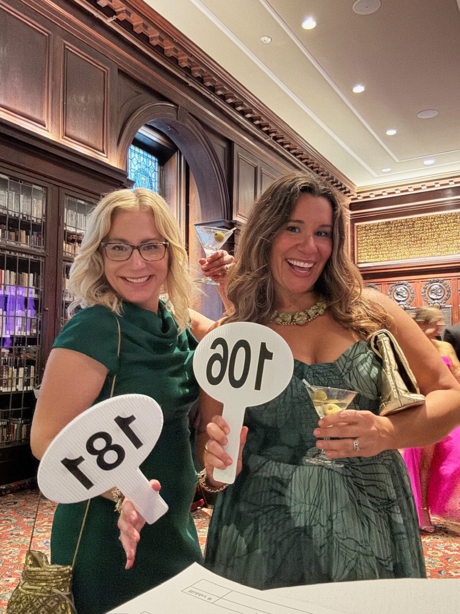 Two women smiling and holding signs with numbers, in an elegant room with wooden shelves and warm lighting.
