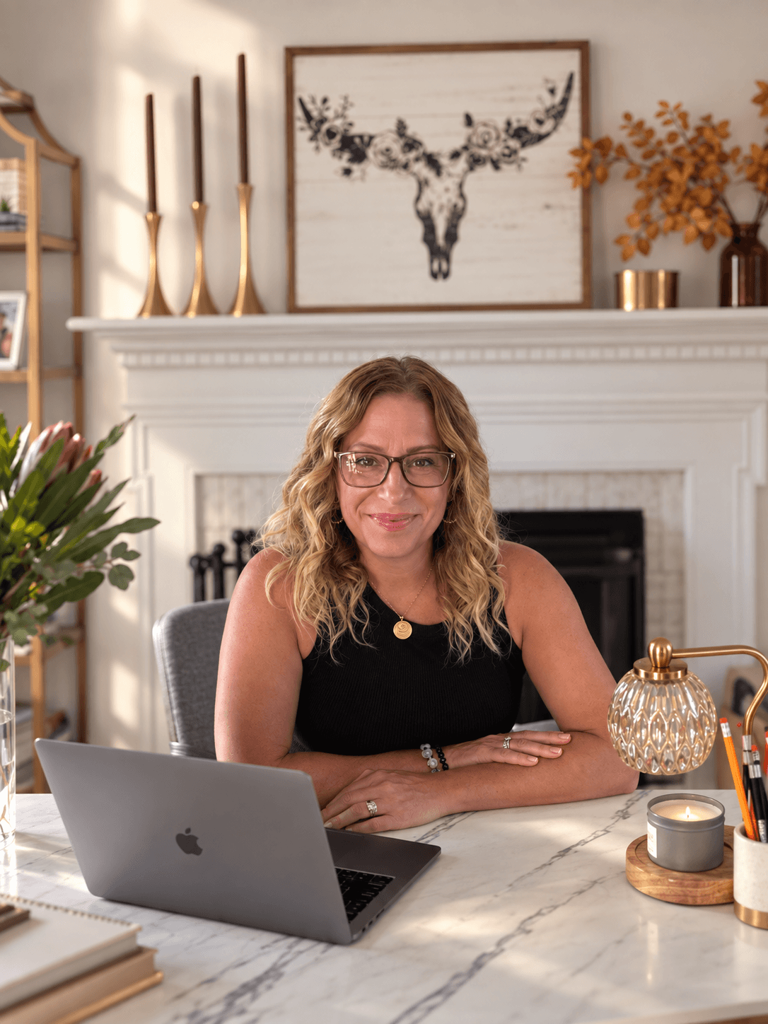 Woman with glasses and wavy hair sitting at a desk with a laptop, in a room with a fireplace and decorative items.