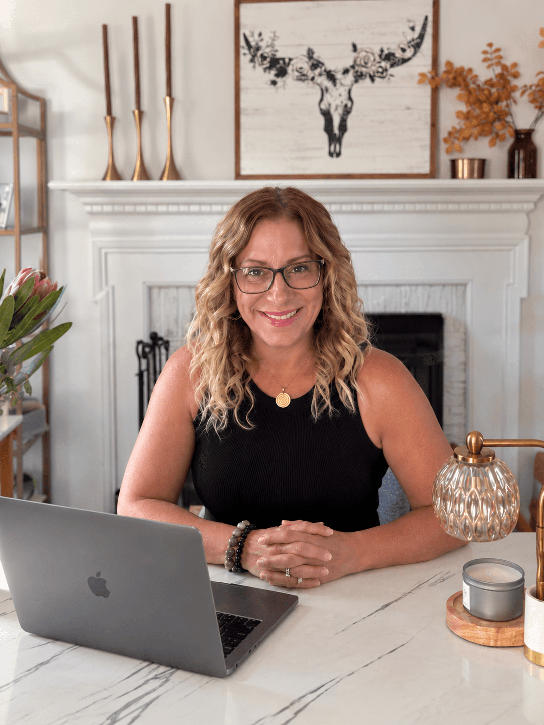 Woman with curly hair and glasses sitting at a desk in front of a fireplace, smiling, with a laptop and decorative items.